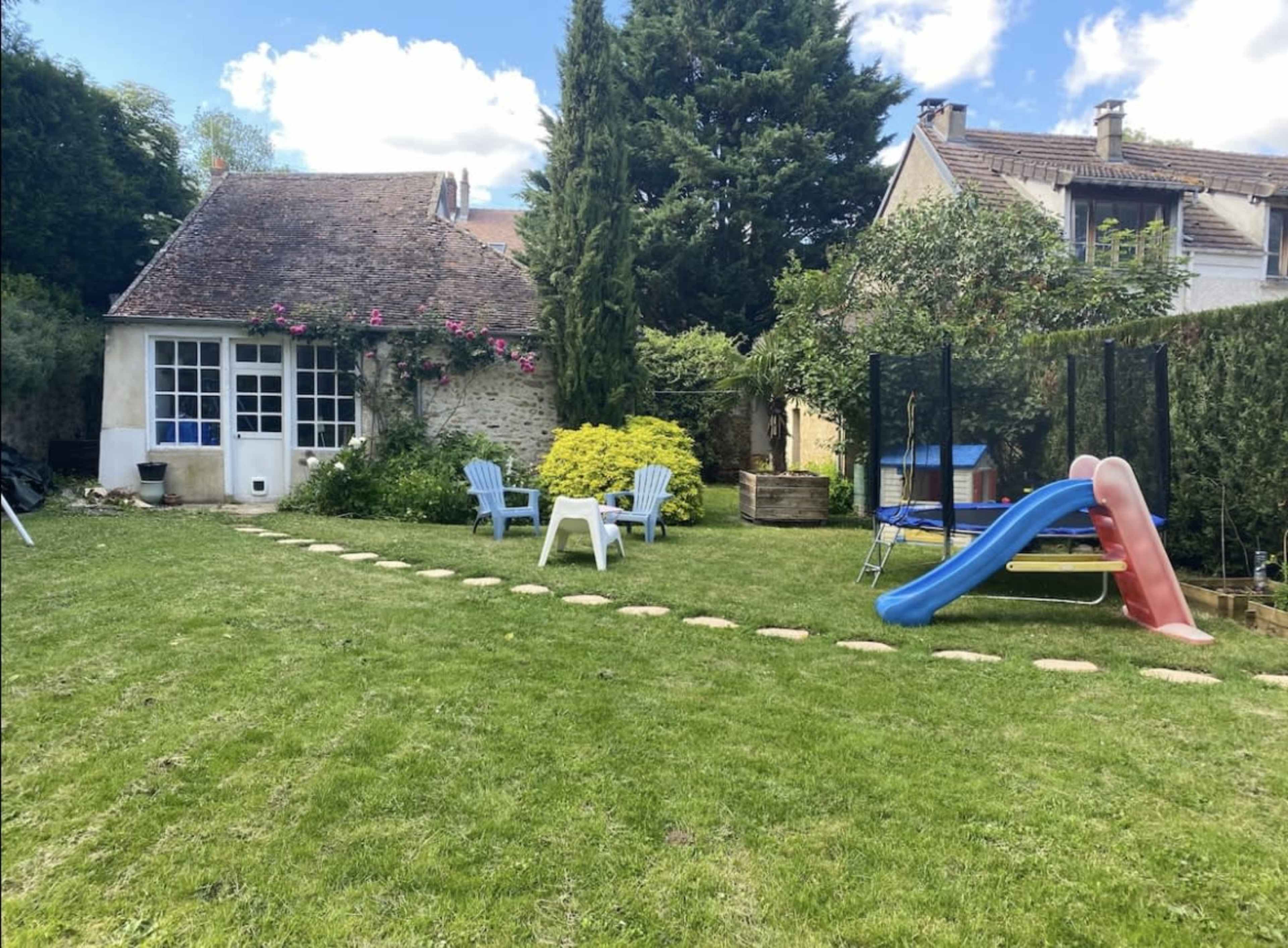 A backyard features a small house with a patio area, a slide, and stone path leading through the grassy yard.