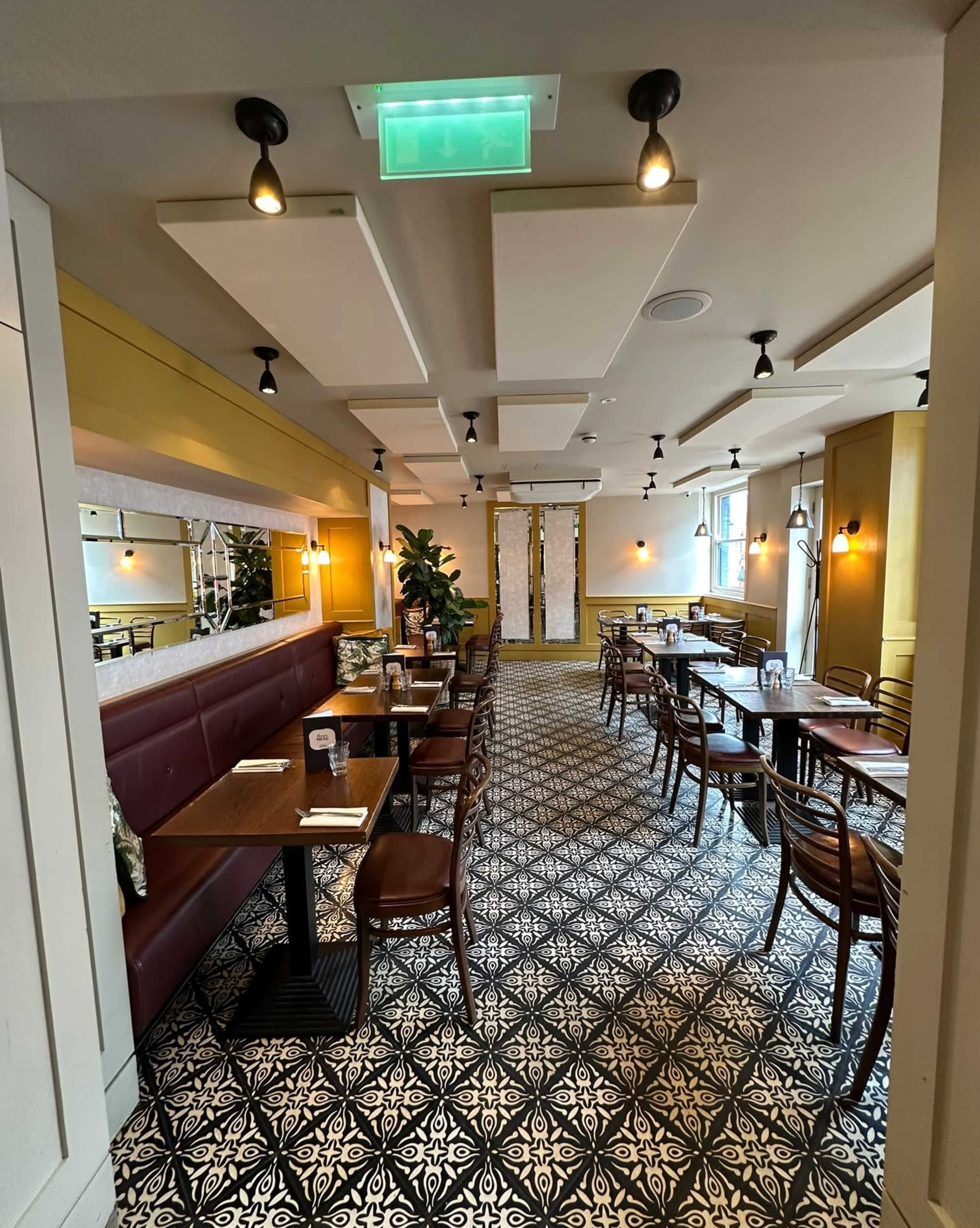 The image shows an interior view of a restaurant featuring a patterned floor, wooden tables, and seating along the walls.