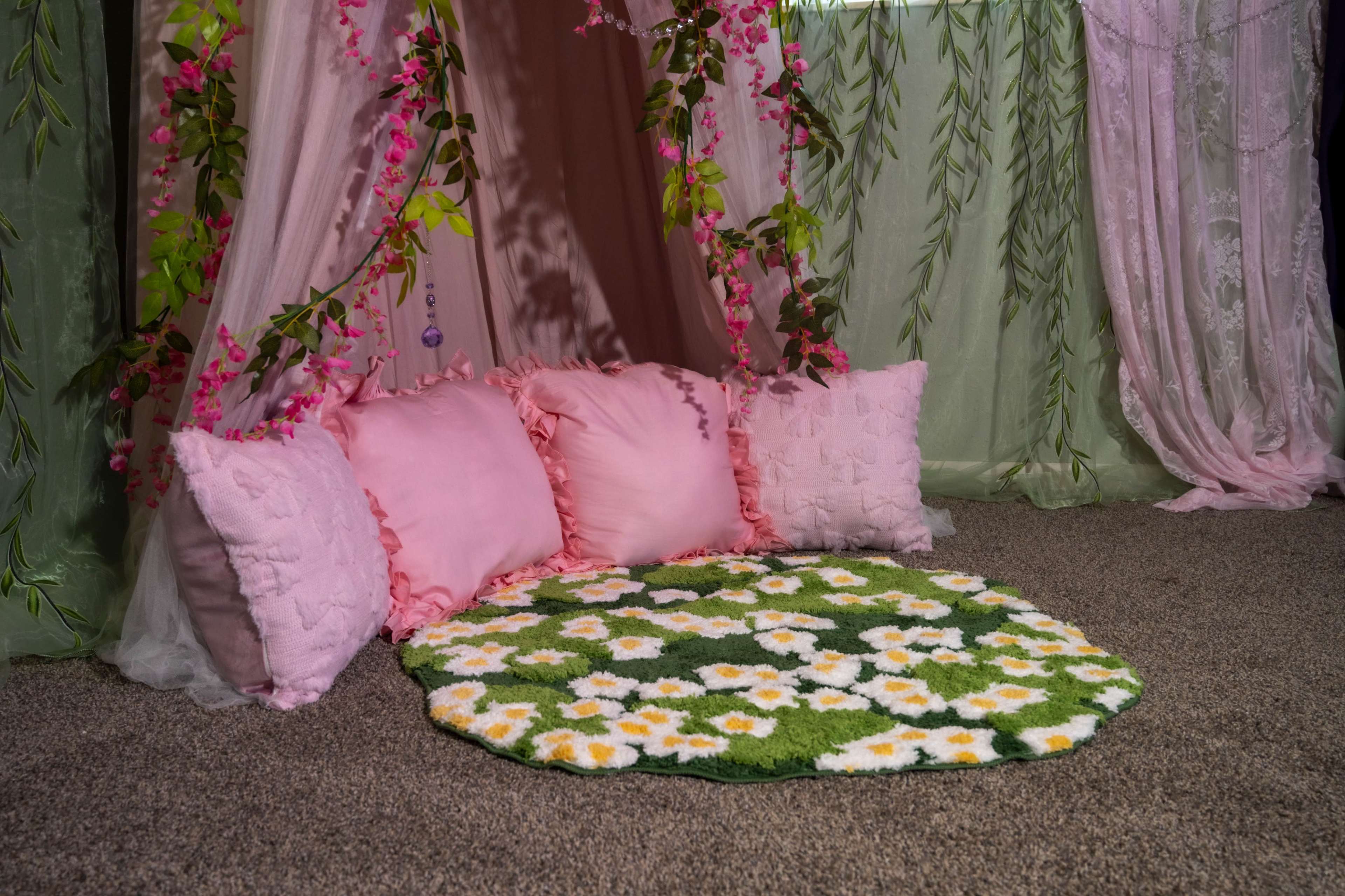 The scene features a circular green rug with white and yellow flower patterns, surrounded by three pink pillows and draped fabric in soft hues, creating a cozy corner.
