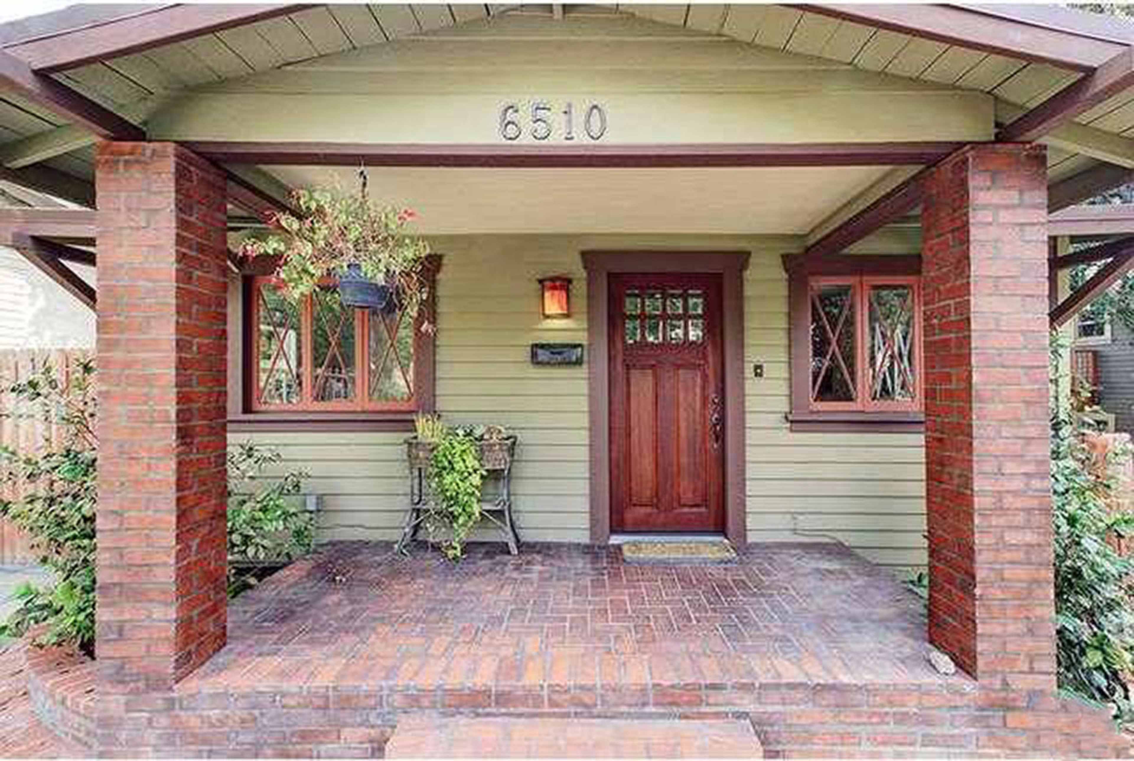 A front porch of a green bungalow with red brick pillars and a wooden door, featuring potted plants and a welcome mat.