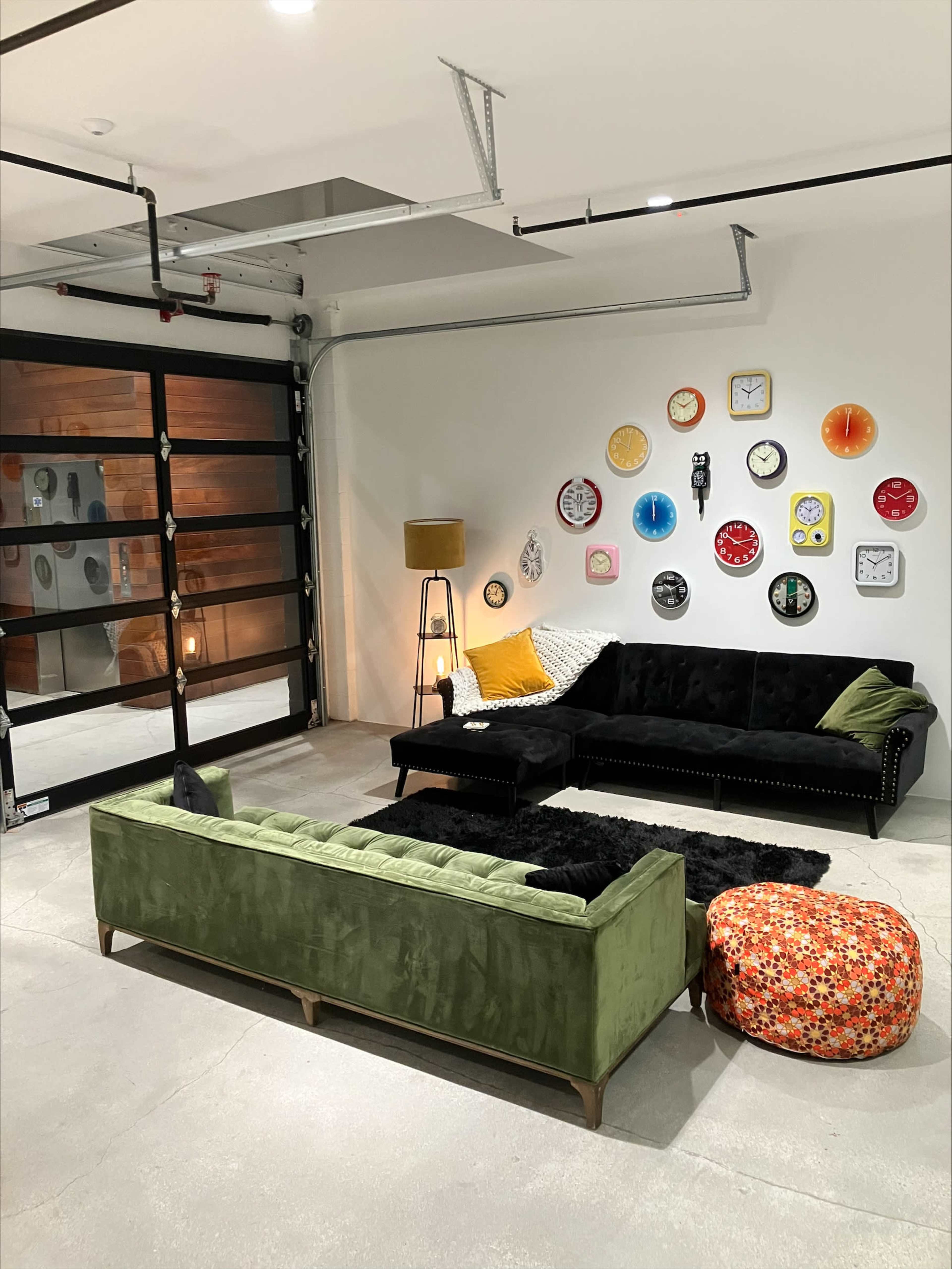 The scene features a modern garage space with two couches, a decorative wall of various clocks, a floor lamp, and an orange patterned ottoman.