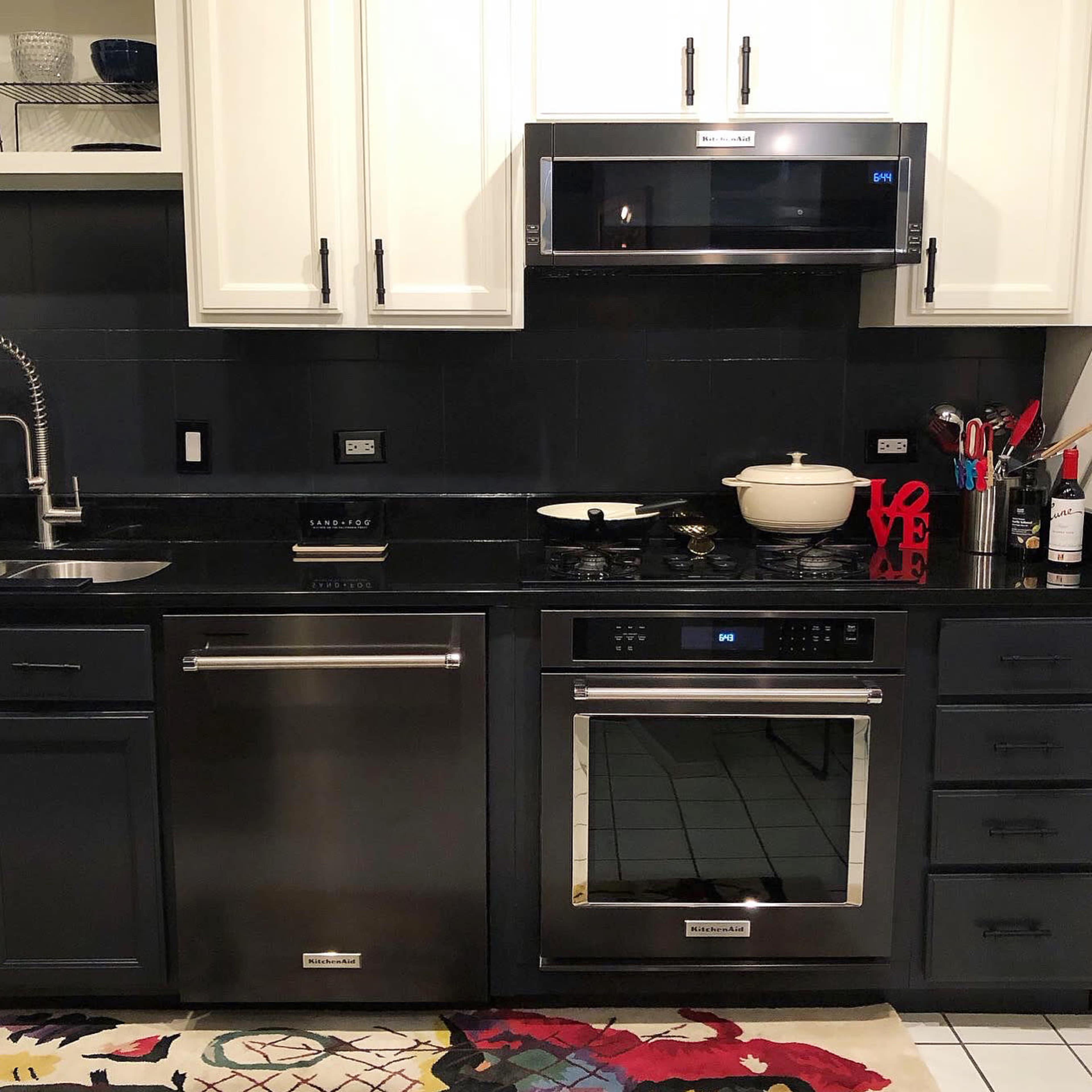 The image shows a modern kitchen with black cabinetry, stainless steel appliances, a sink, and a colorful rug on the floor.