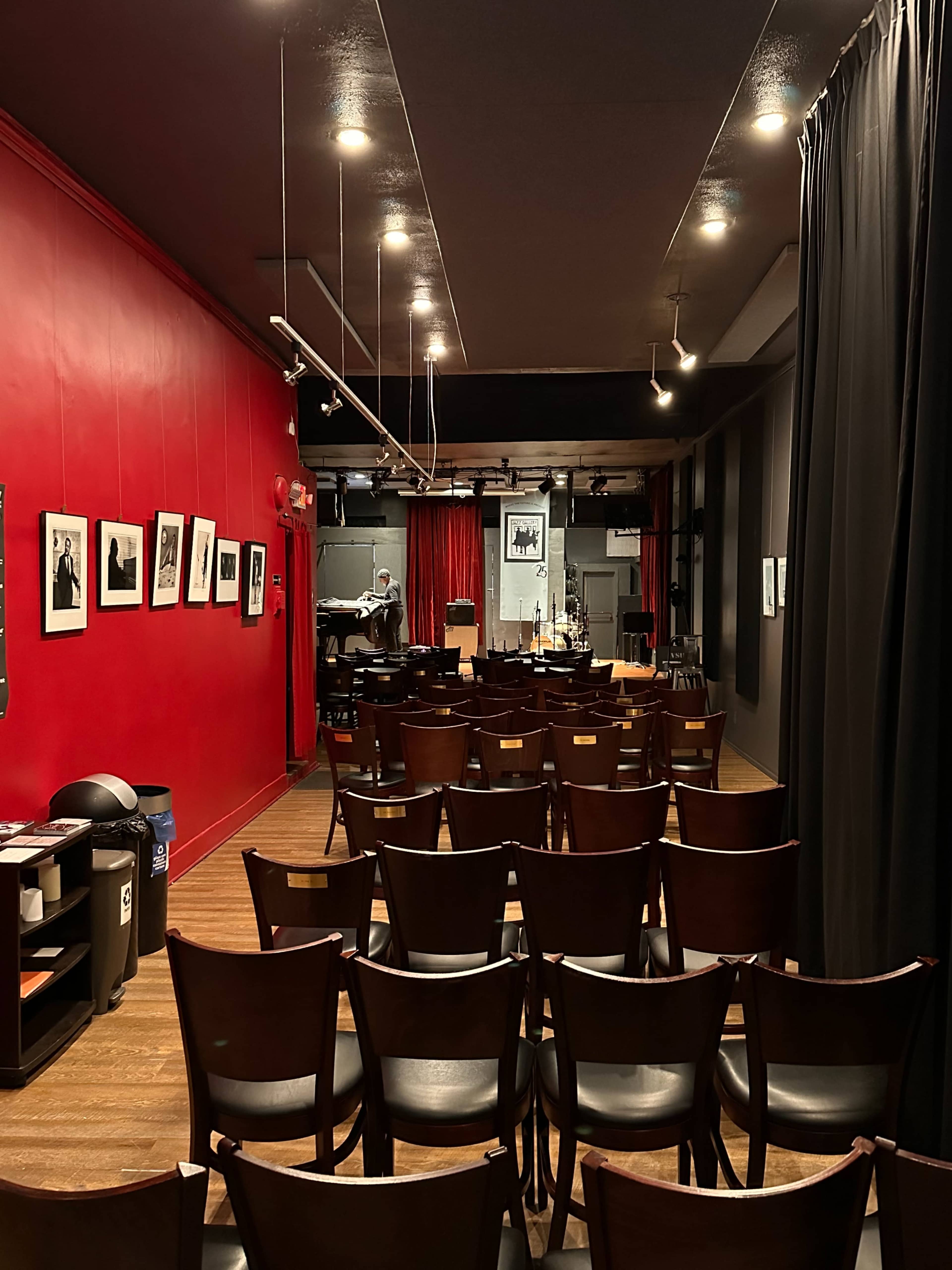 The image shows a row of wooden chairs set up in a dimly lit performance space with red walls, framed photographs, and a small stage in the background.