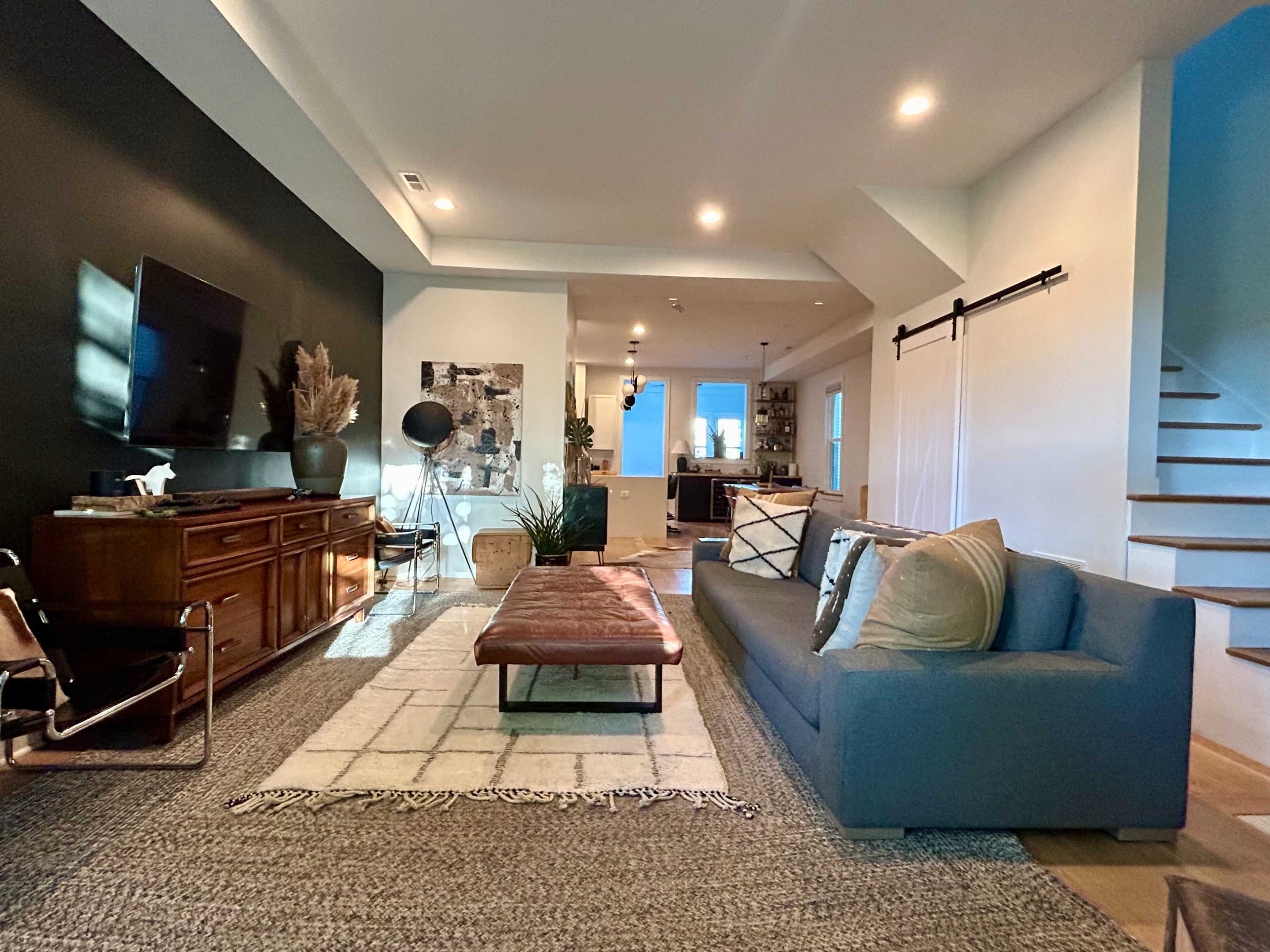 The image shows a modern living room with a sectional sofa, a leather ottoman, a large area rug, and a staircase in the background, featuring a mix of decor elements against contrasting wall colors.