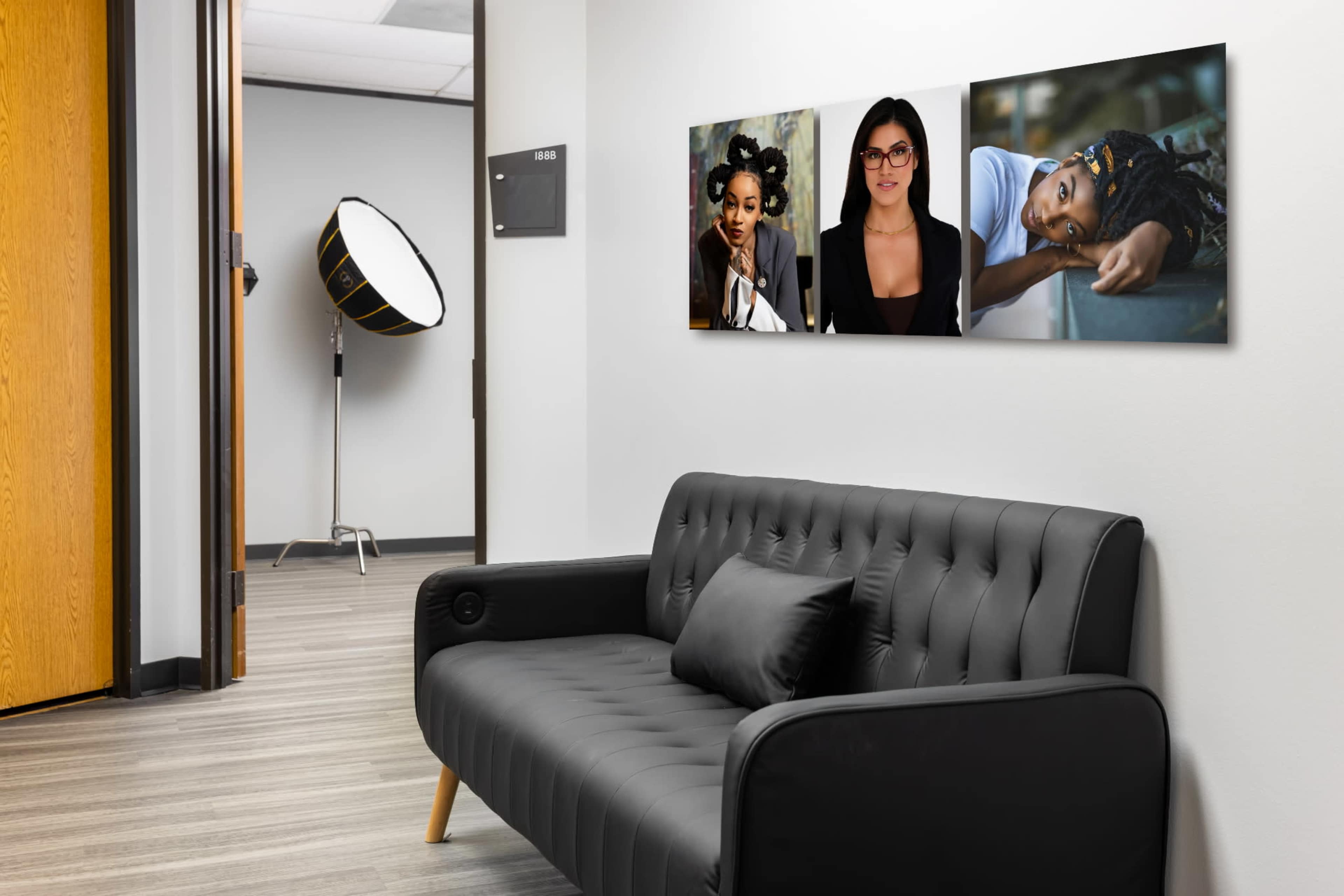 A black tufted sofa is positioned against a wall adorned with two framed portraits, while a large softbox light stands nearby in a well-lit office space.