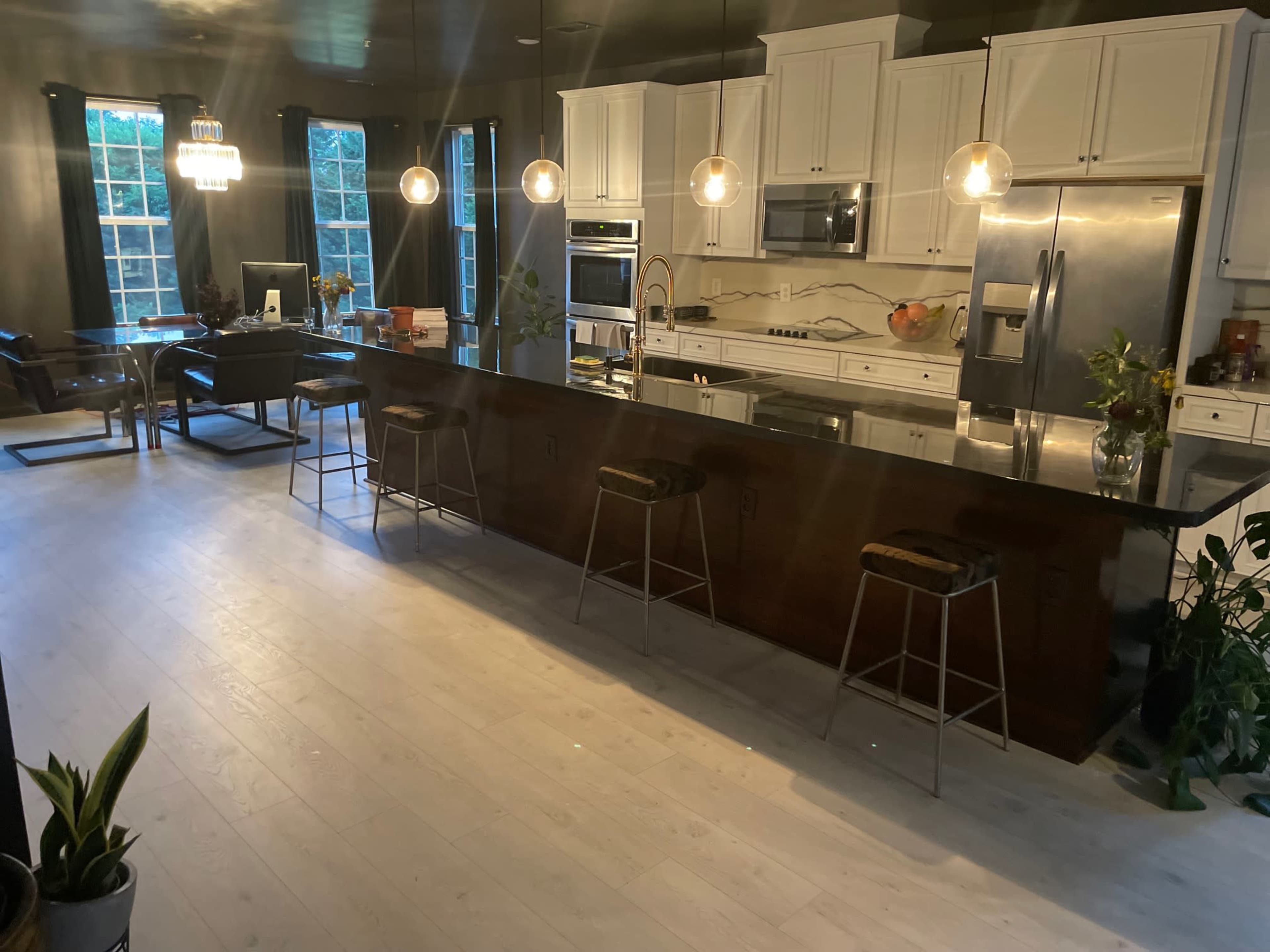 A modern kitchen features white cabinetry, a large island with bar stools, and pendant lighting, illuminated by natural light from large windows.