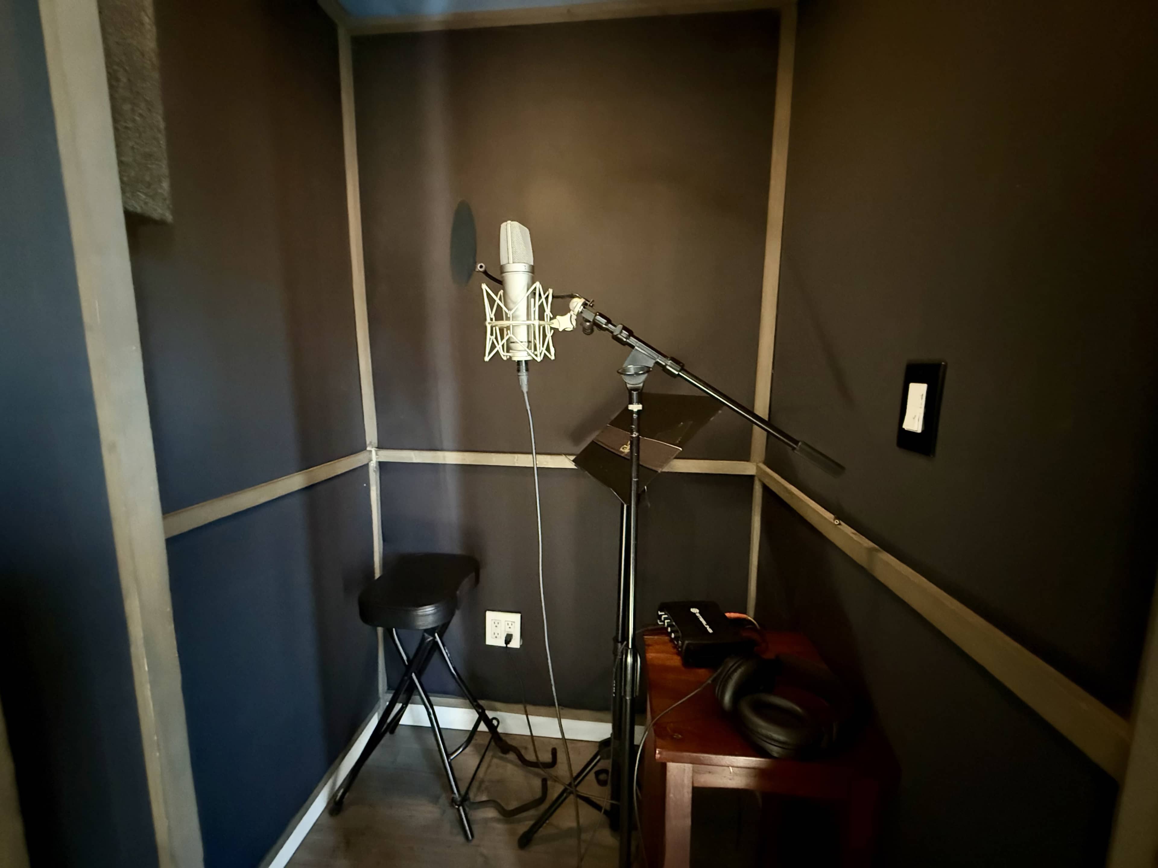 The image shows a small recording booth with a microphone on a stand, a stool, a music stand, and equipment on a table.