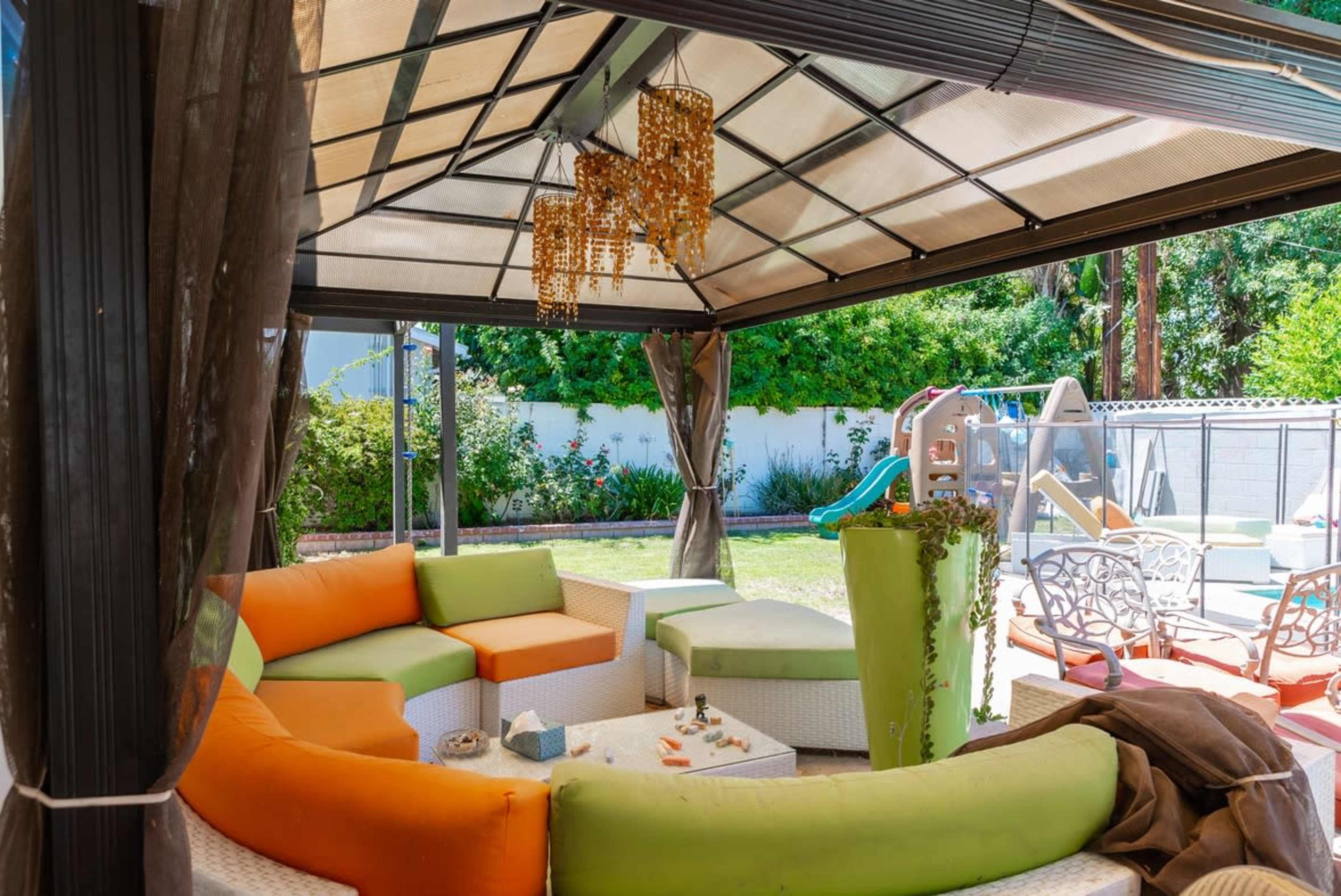A covered outdoor seating area features curved, colorful cushions and a chandelier, overlooking a lawn with playground equipment.