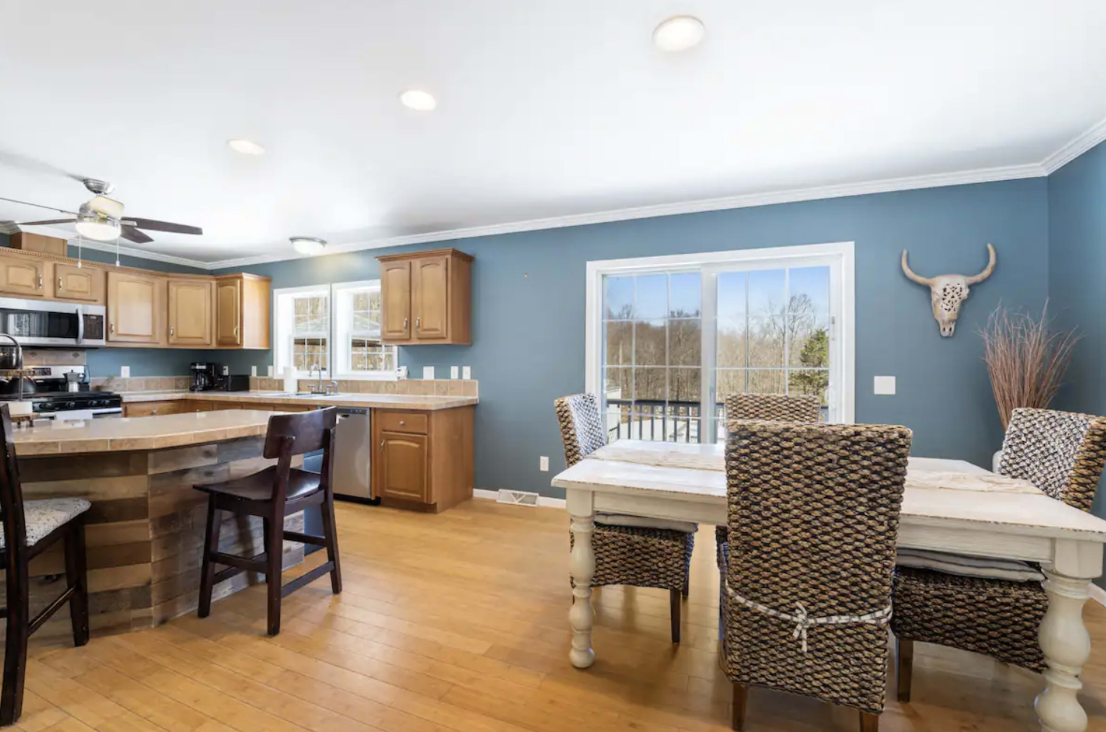The image shows a kitchen area with wooden cabinets, a dining table with woven chairs, and large windows overlooking a natural landscape.