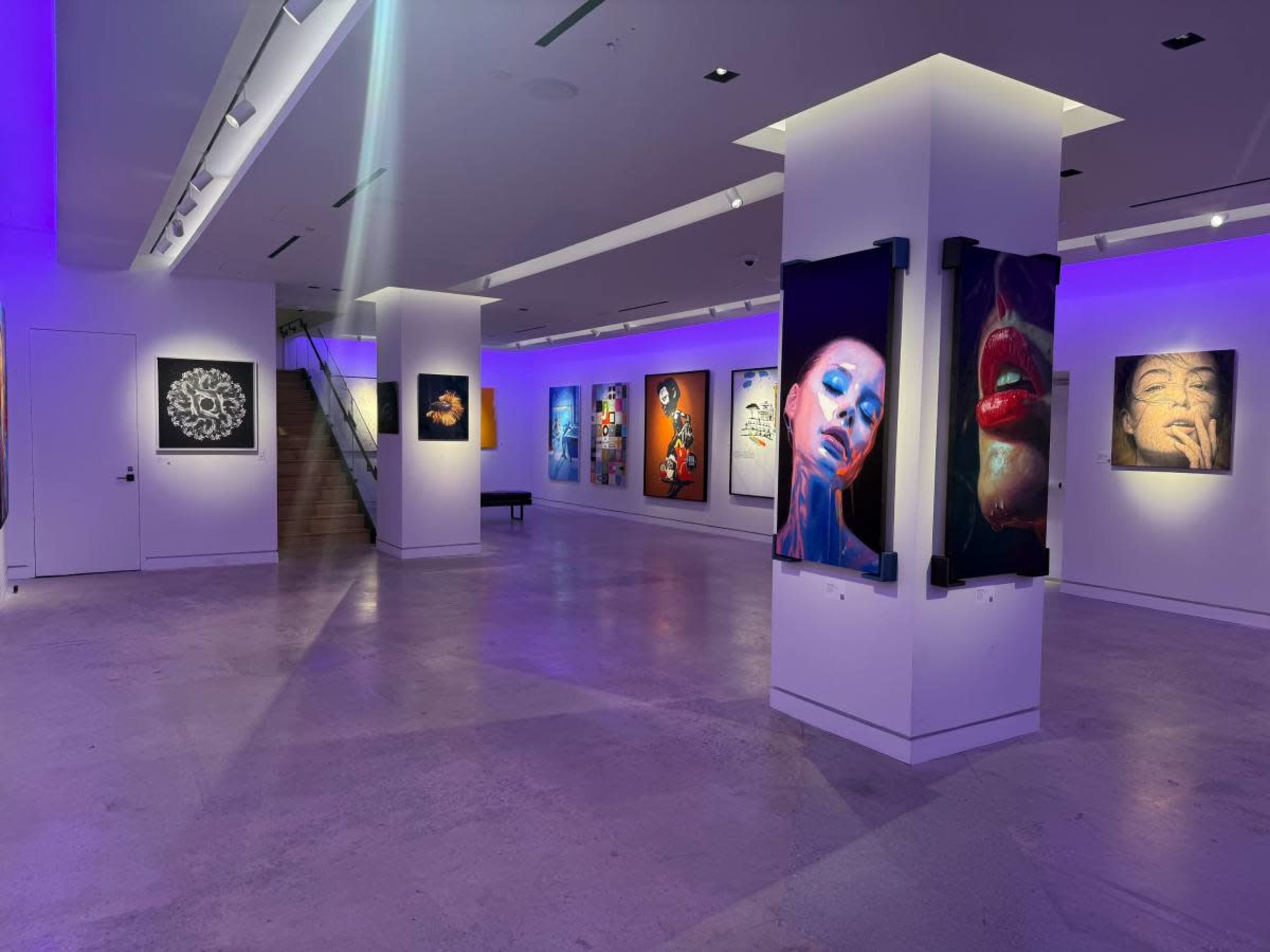 The image shows an art gallery with illuminated walls displaying various framed artworks and a staircase in the background.