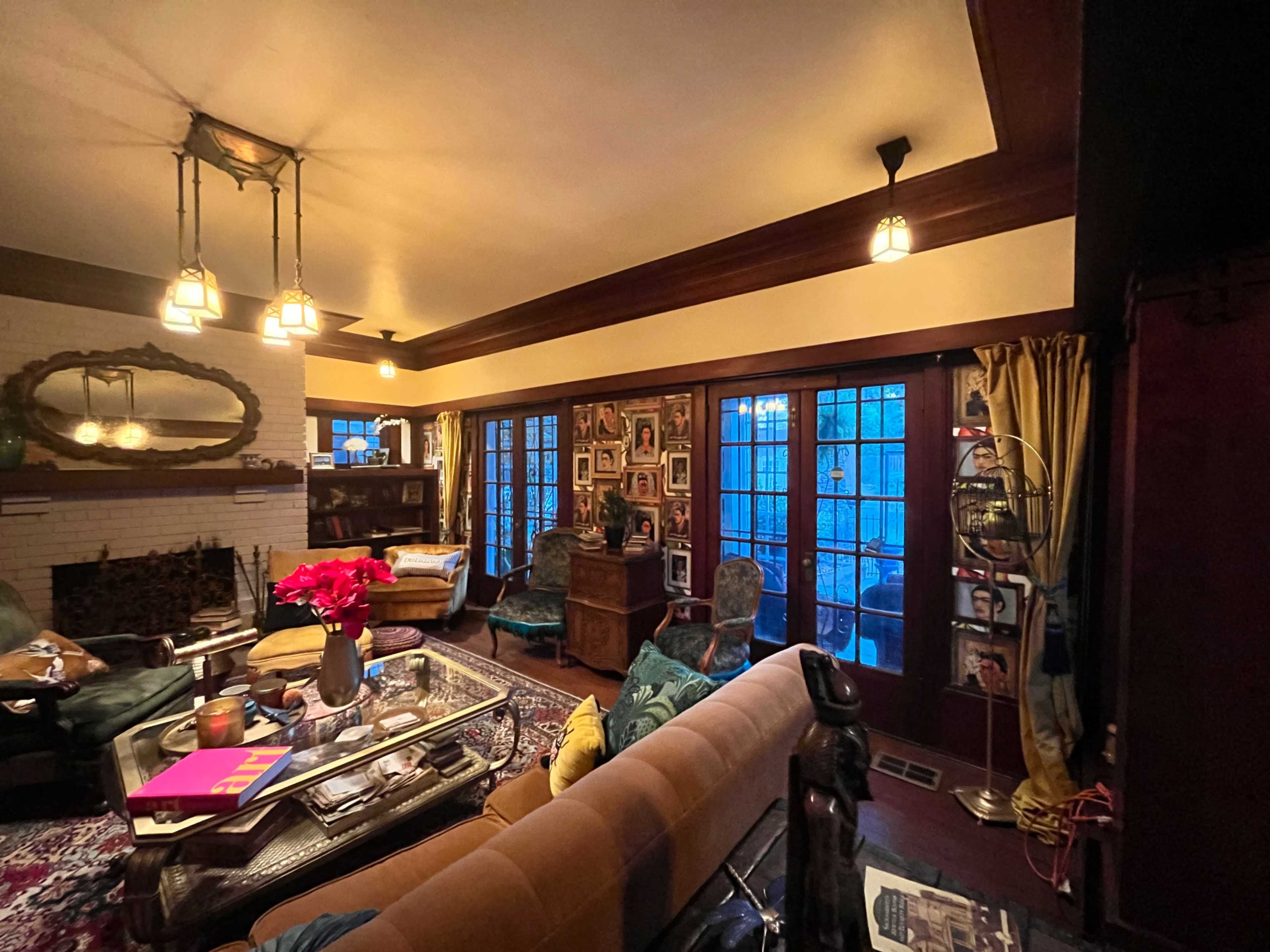 The image shows a cozy living room with a mix of vintage furniture, a coffee table adorned with books and flowers, and large windows displaying an evening blue light.