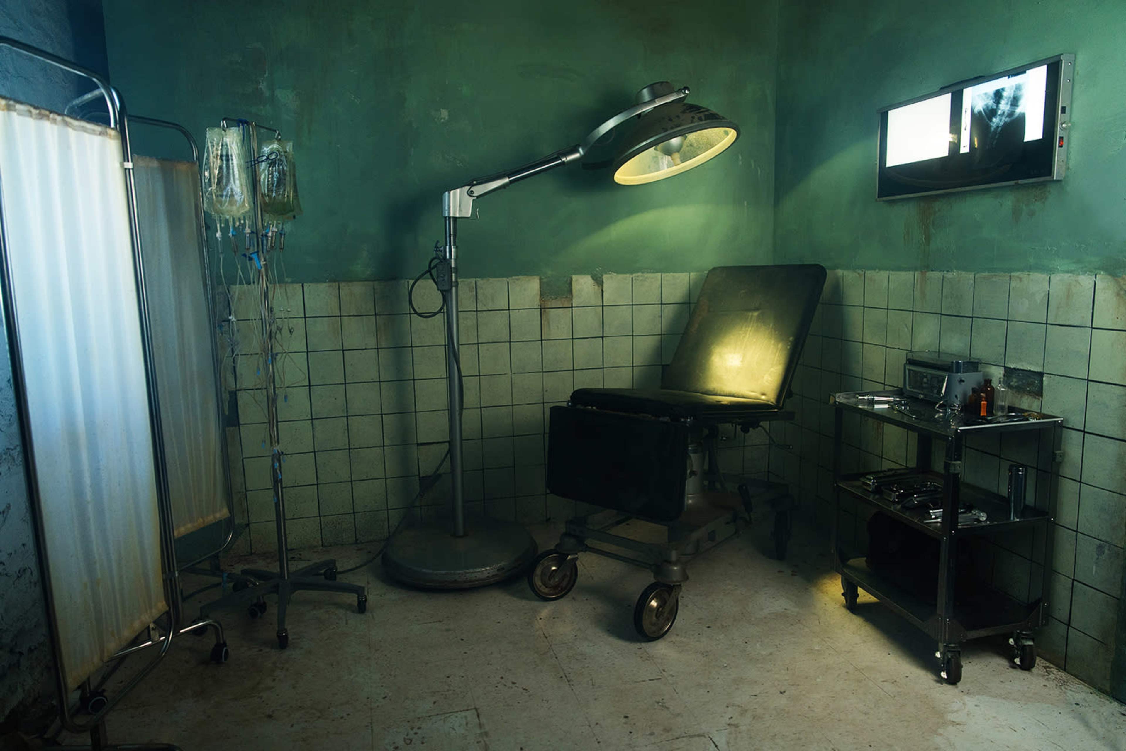 The image shows an empty, dimly lit medical room with a retractable examination table, an IV stand, and a small cart holding various medical equipment.