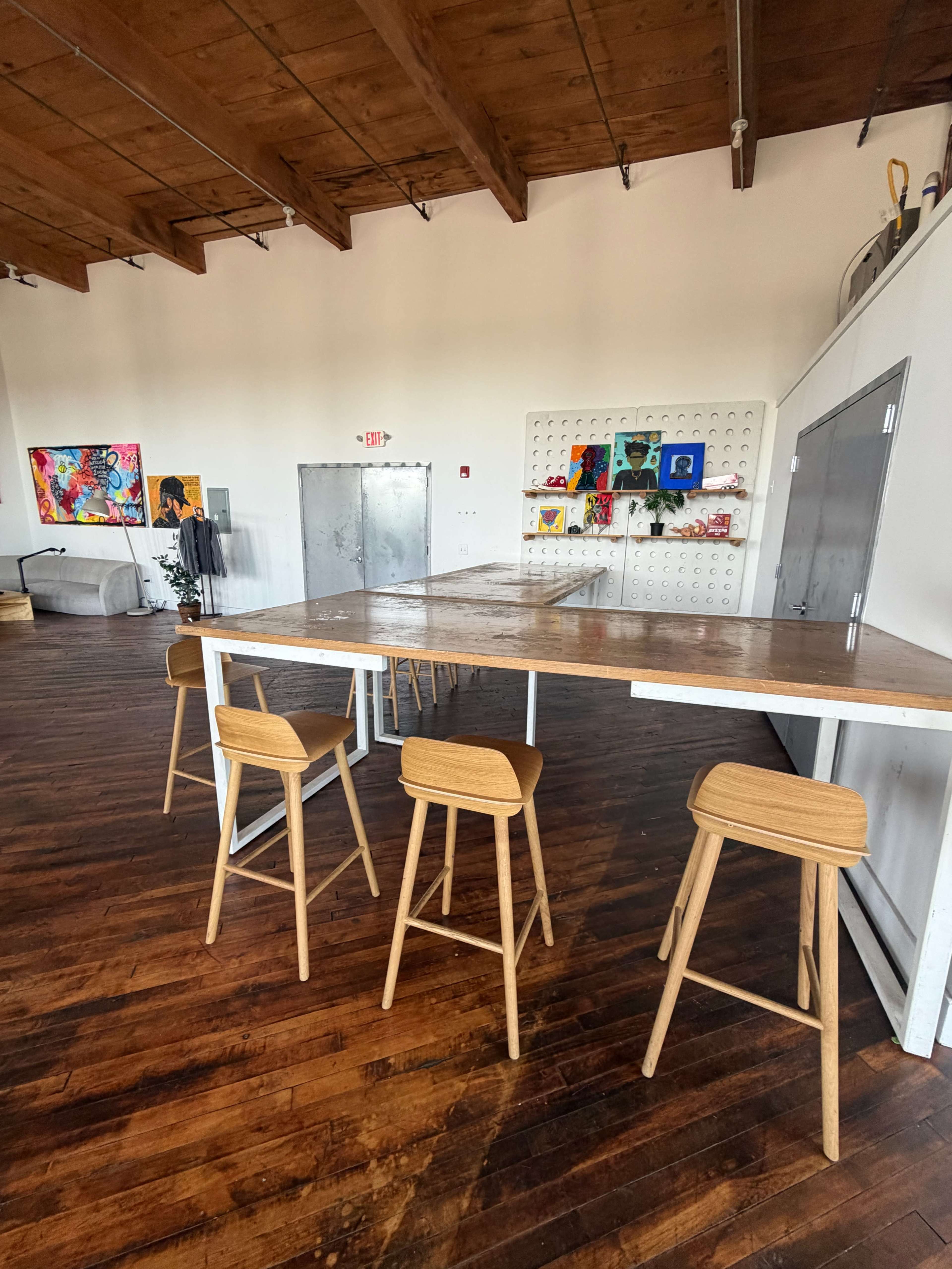The image shows a spacious interior with wooden flooring, a large table surrounded by several stools, and colorful artwork displayed on the walls.