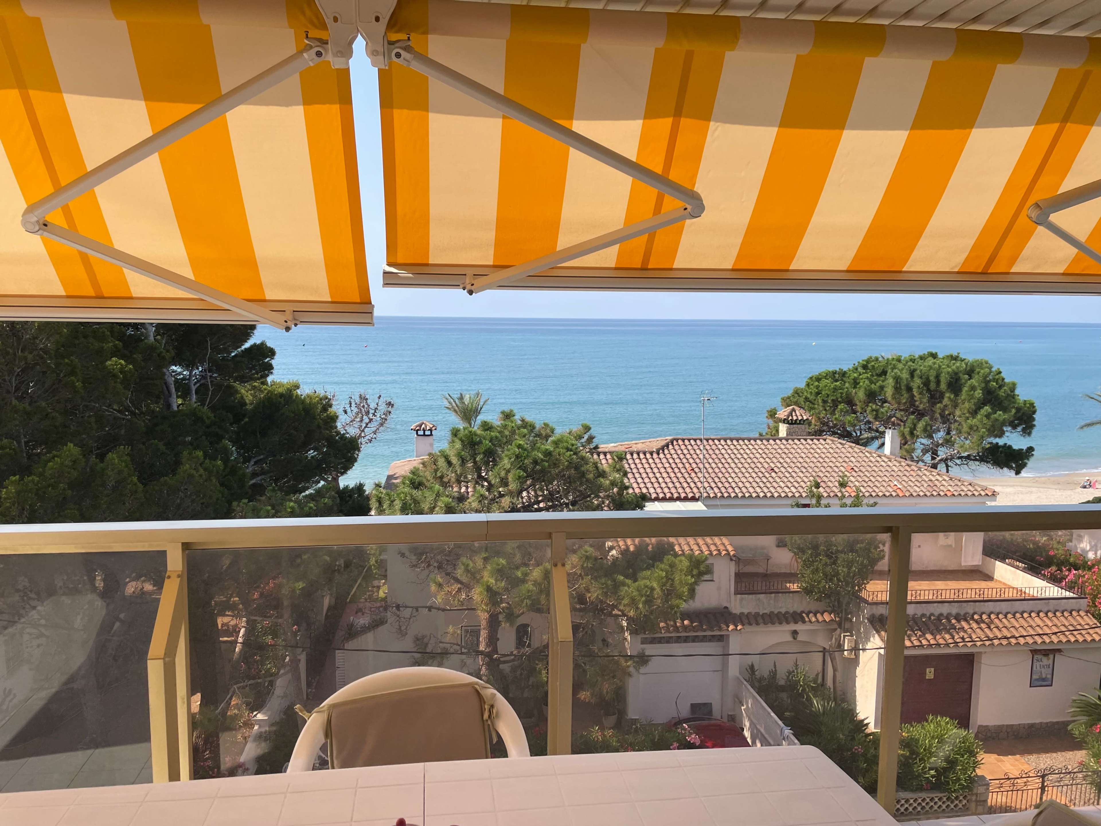 The image shows a balcony with yellow and white striped awnings overlooking a calm beach and coastal properties.