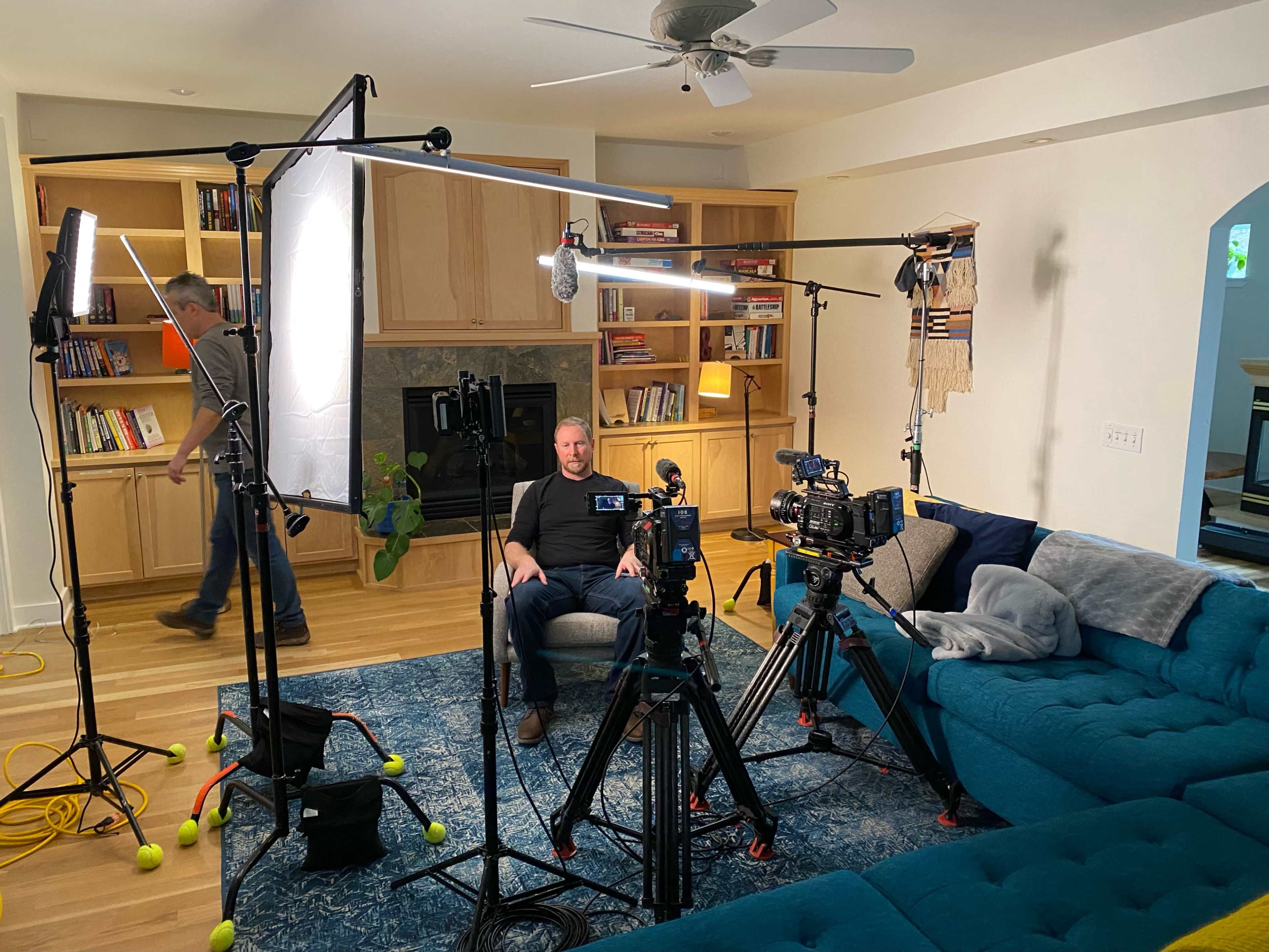 The scene shows a living room set up for a video shoot, featuring multiple cameras, lights, and a person sitting in a chair amidst a backdrop of bookshelves.