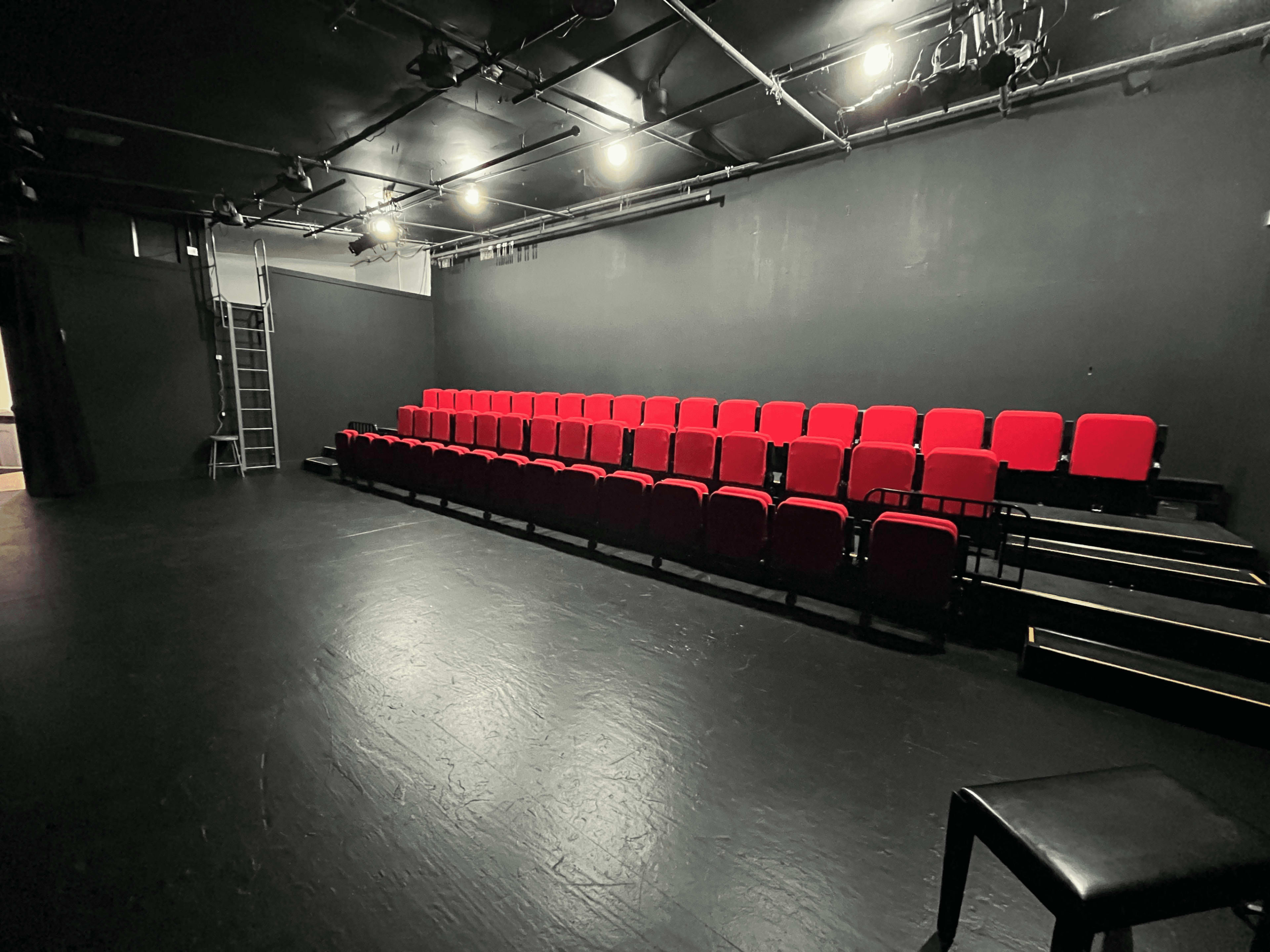 The image shows an empty black box theater with rows of red seats arranged in a staggered formation.