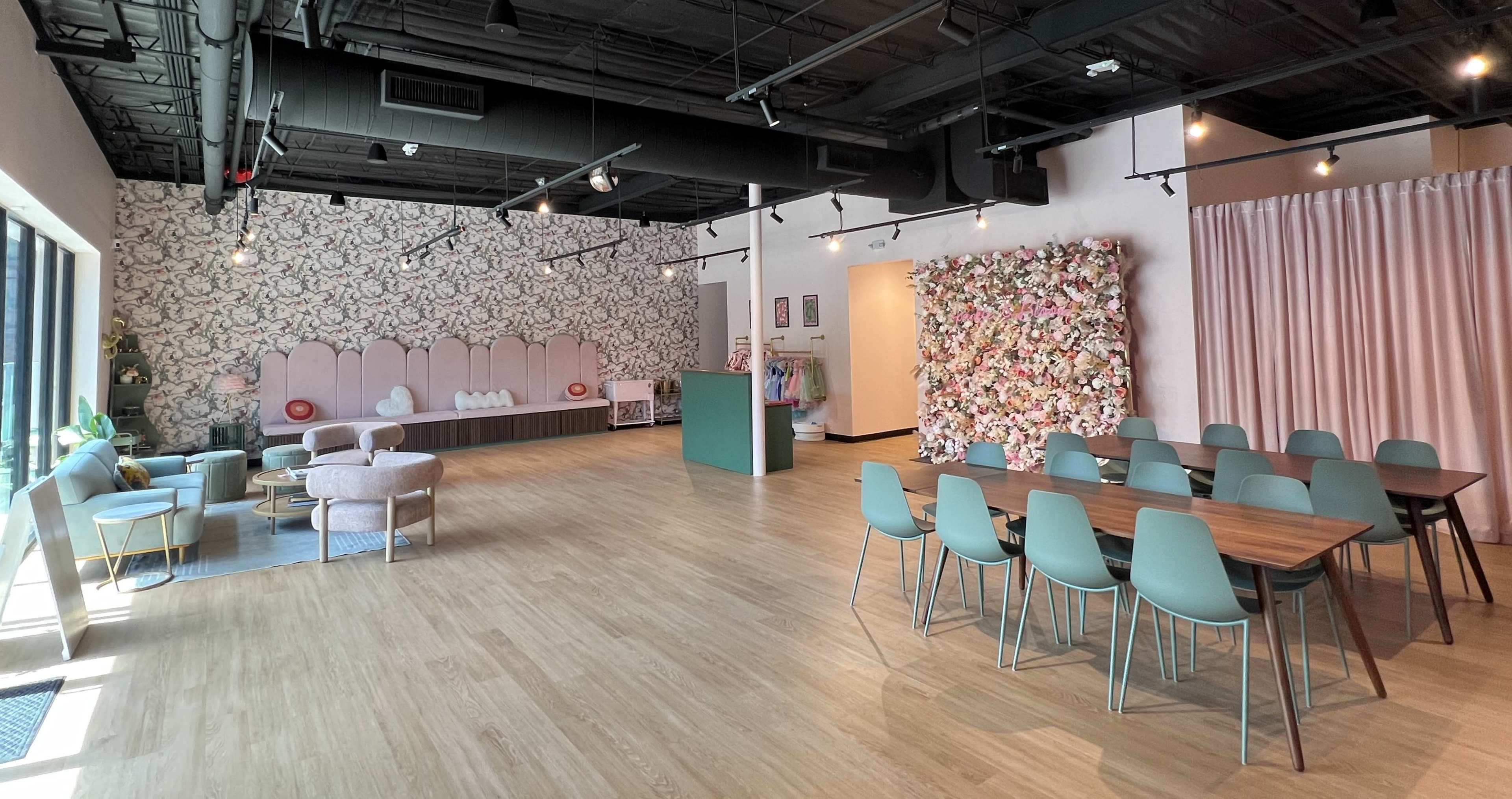 The image shows a spacious interior of a modern venue featuring a floral wall, a seating area with plush chairs, and a large table with chairs arranged for group gatherings.