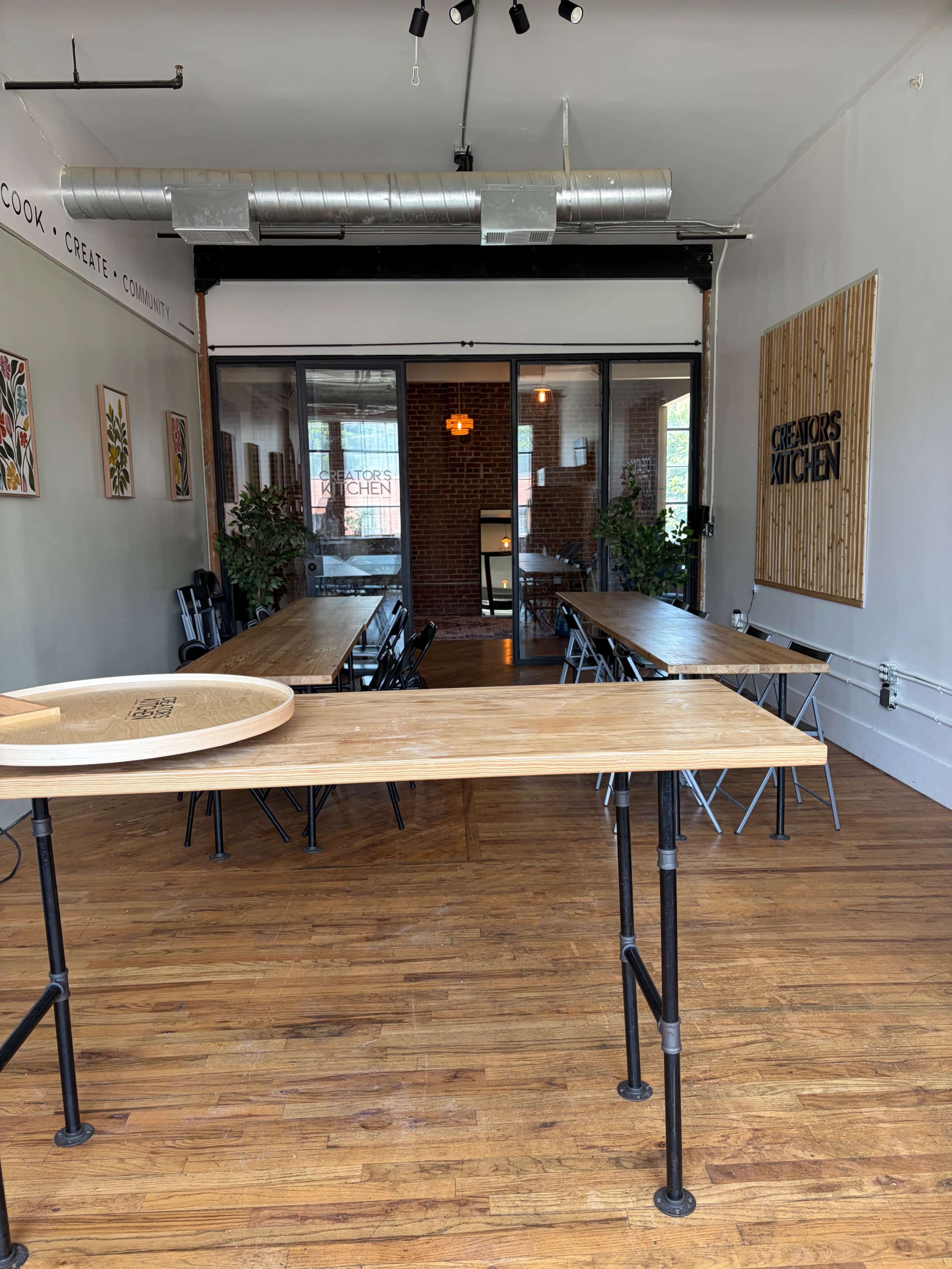 The image shows a spacious, industrial-style kitchen and dining area with wooden tables, black chairs, and large windows letting in natural light.