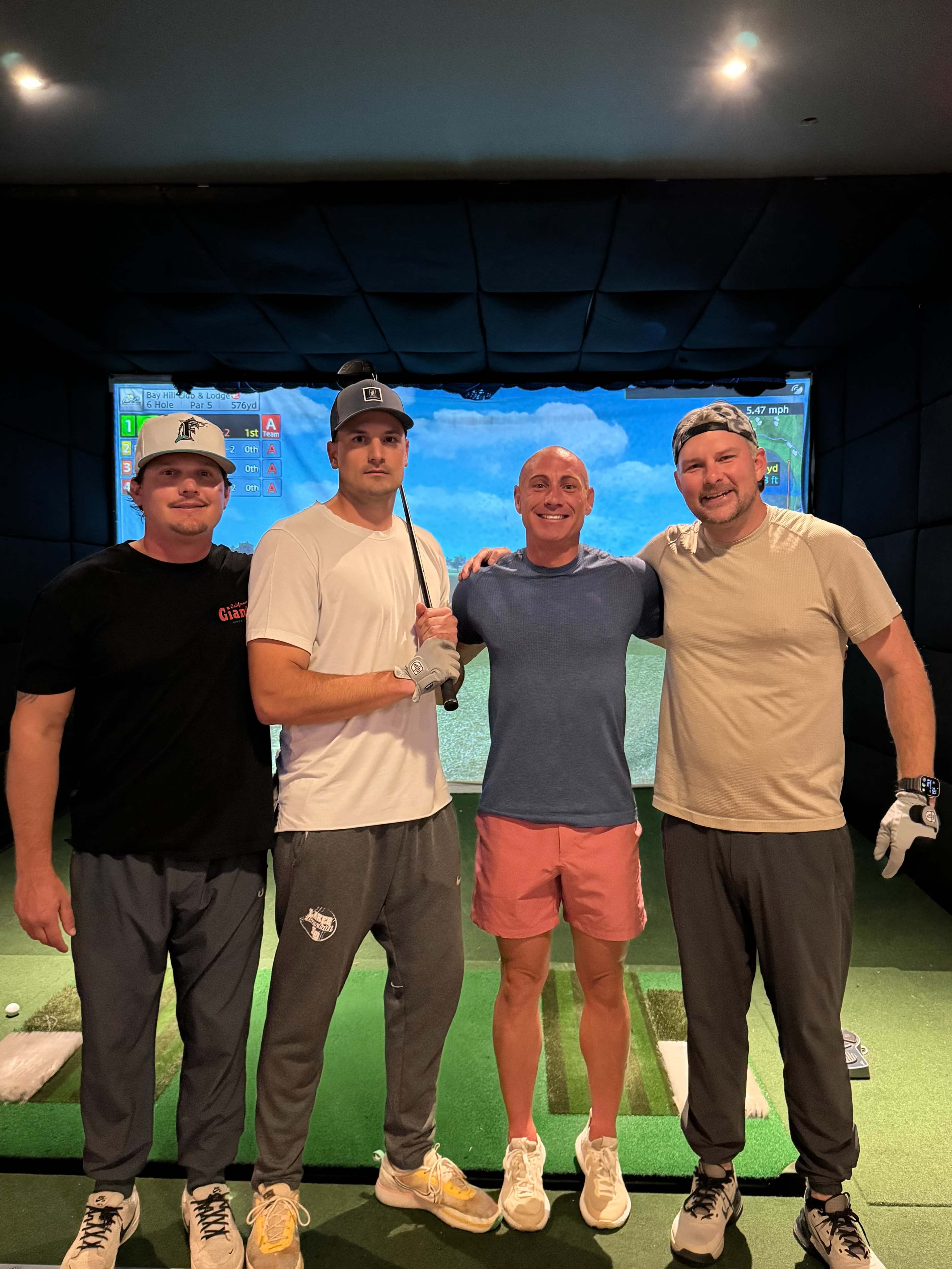 Four men stand inside a golf simulator room, posing together with a digital golf course displayed on a screen behind them.