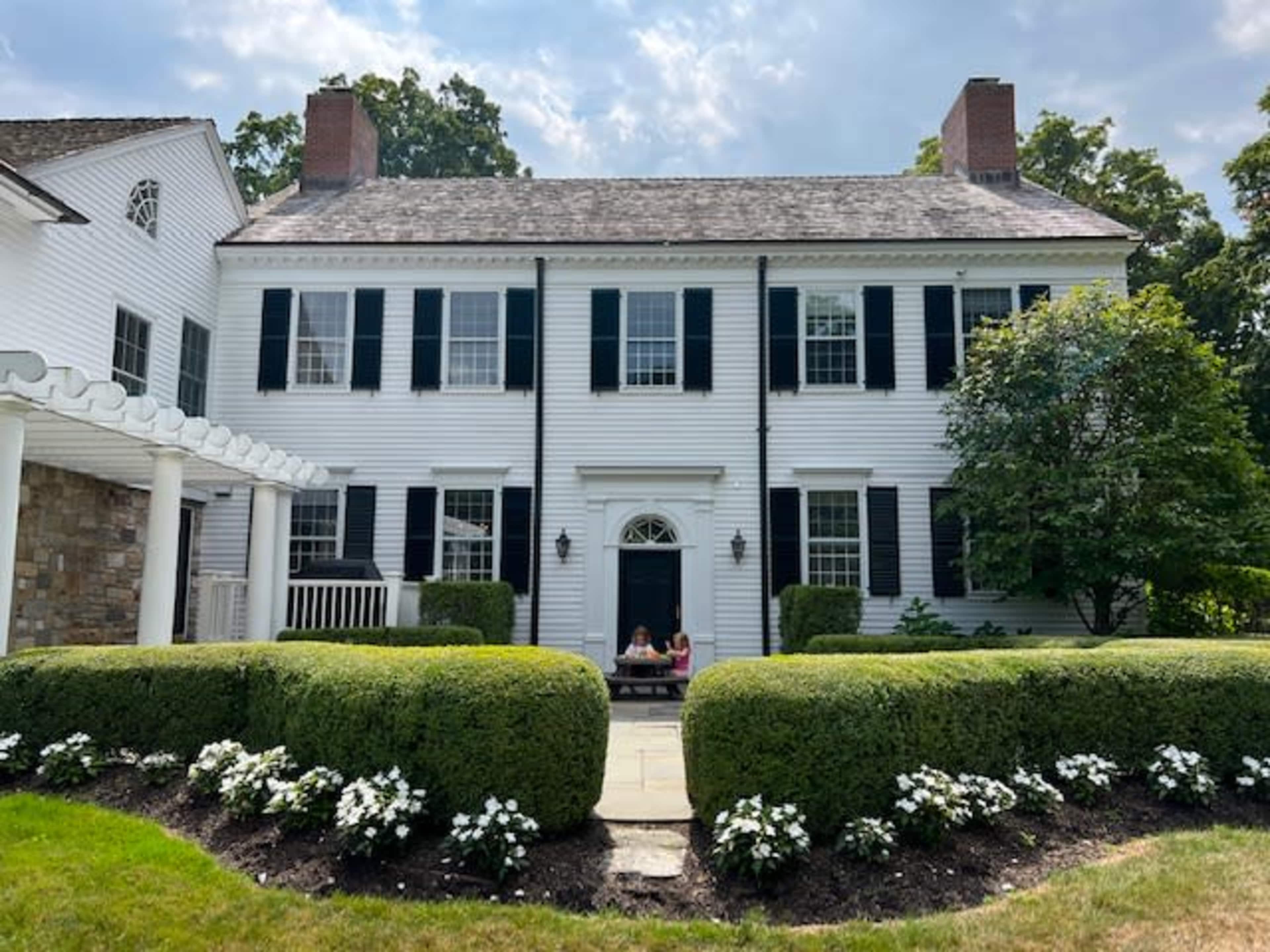 A large white colonial-style house with black shutters is surrounded by meticulously trimmed hedges and flower beds in front.