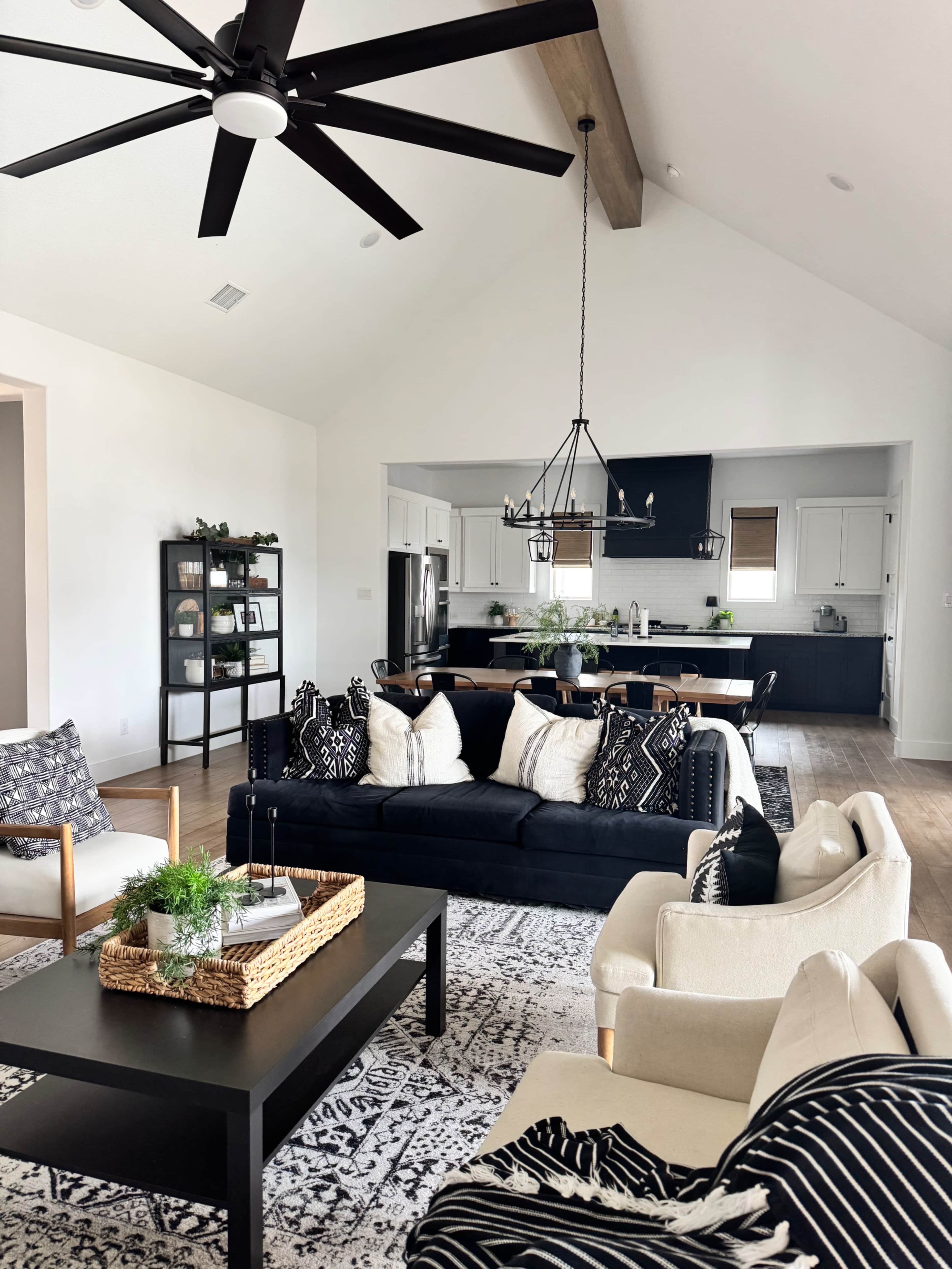 The image shows a modern living room with a black sofa, light-colored chairs, a coffee table, and an open kitchen area in the background featuring dark cabinetry.