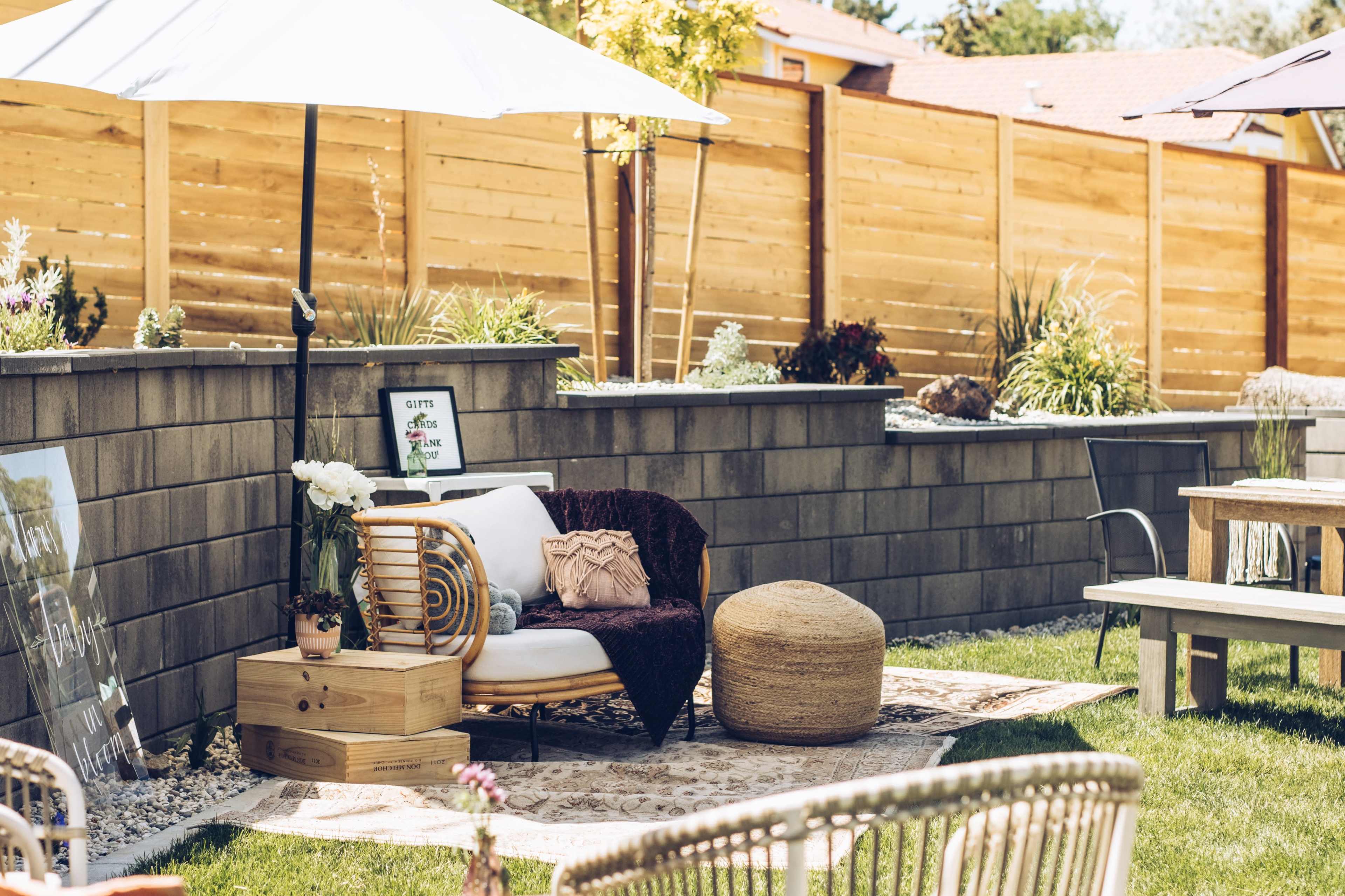 A cozy outdoor seating area features a rattan chair, a round pouf, and decorative plants, set against a wooden fence and grass.