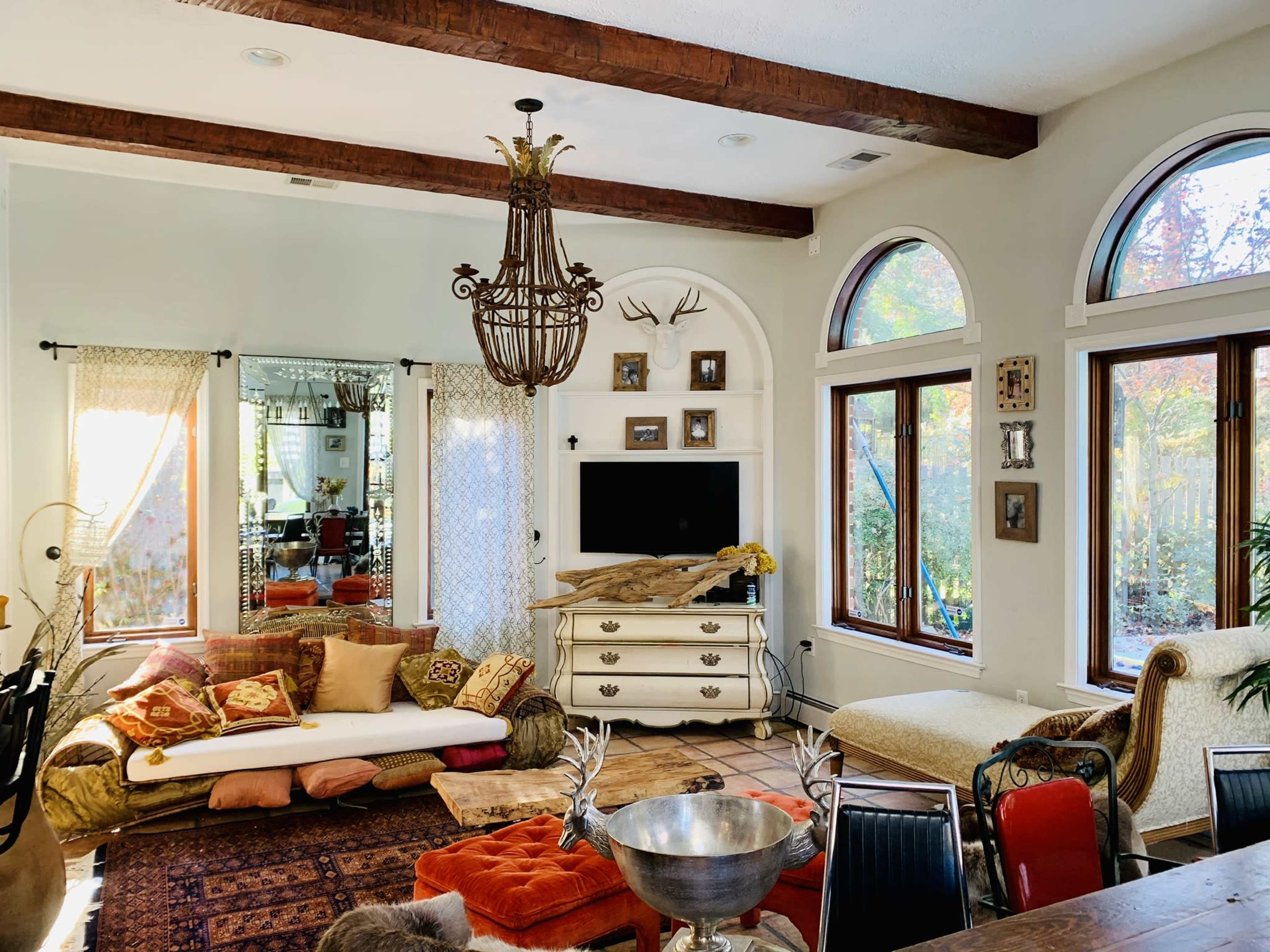 The spacious living room features large windows, a rustic chandelier, a comfortable seating area with decorative pillows, and a central entertainment unit.