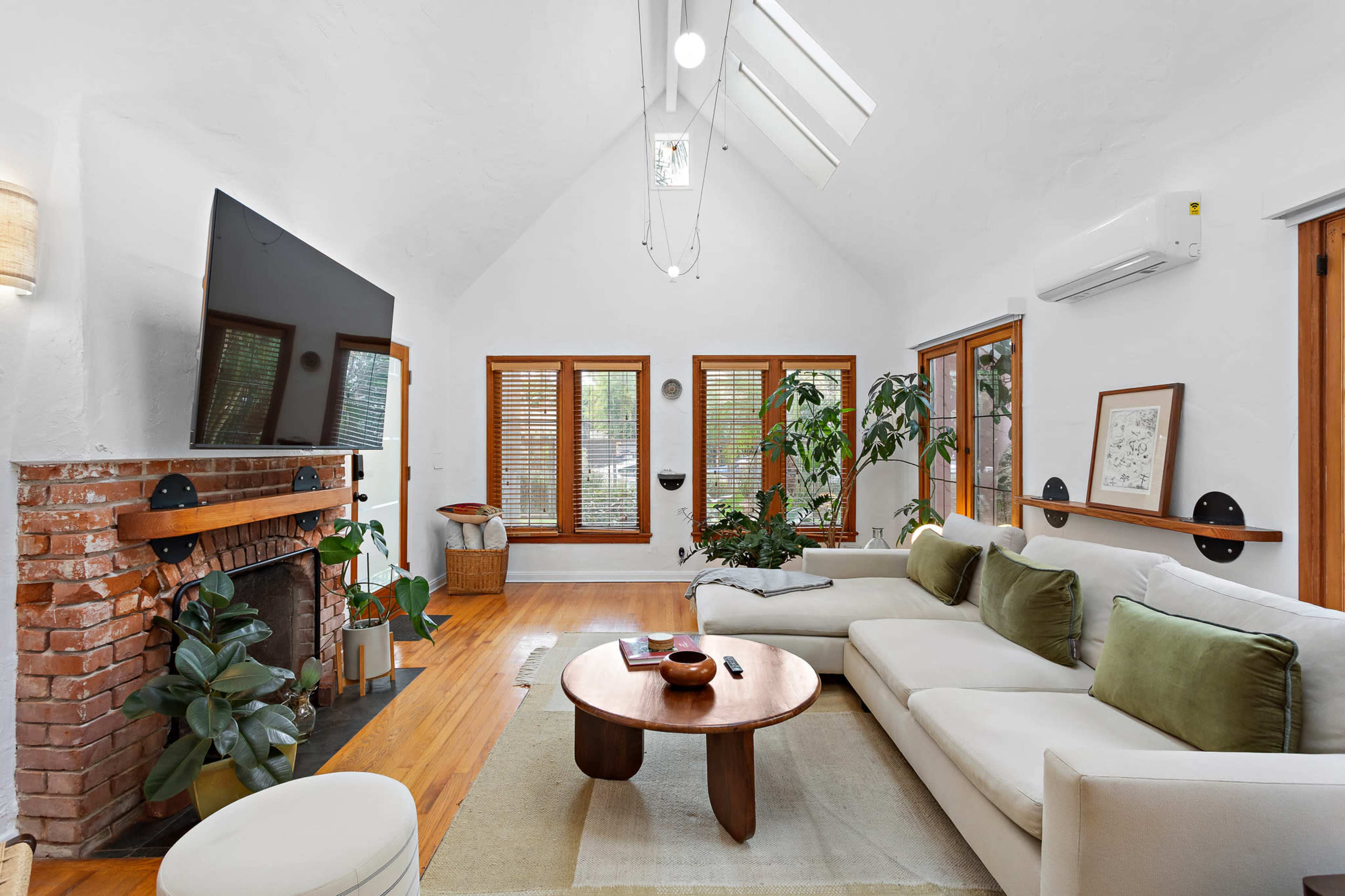The living room features a light-colored sofa, a circular wooden coffee table, and large windows with wooden shutters, complemented by indoor plants and a brick fireplace.