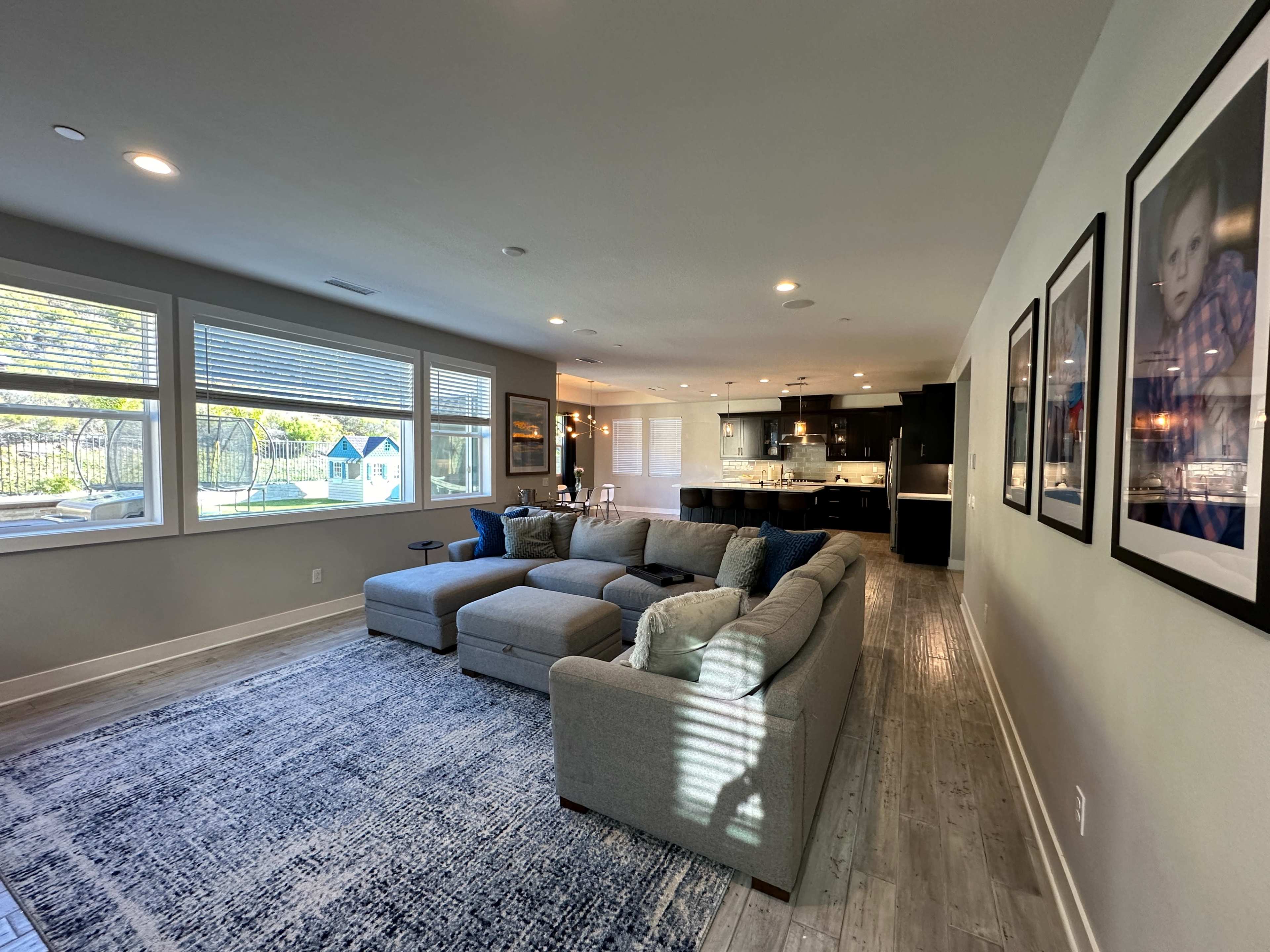 A modern living room featuring a large gray sectional sofa, a patterned area rug, and a kitchen area visible in the background with natural light coming through multiple windows.