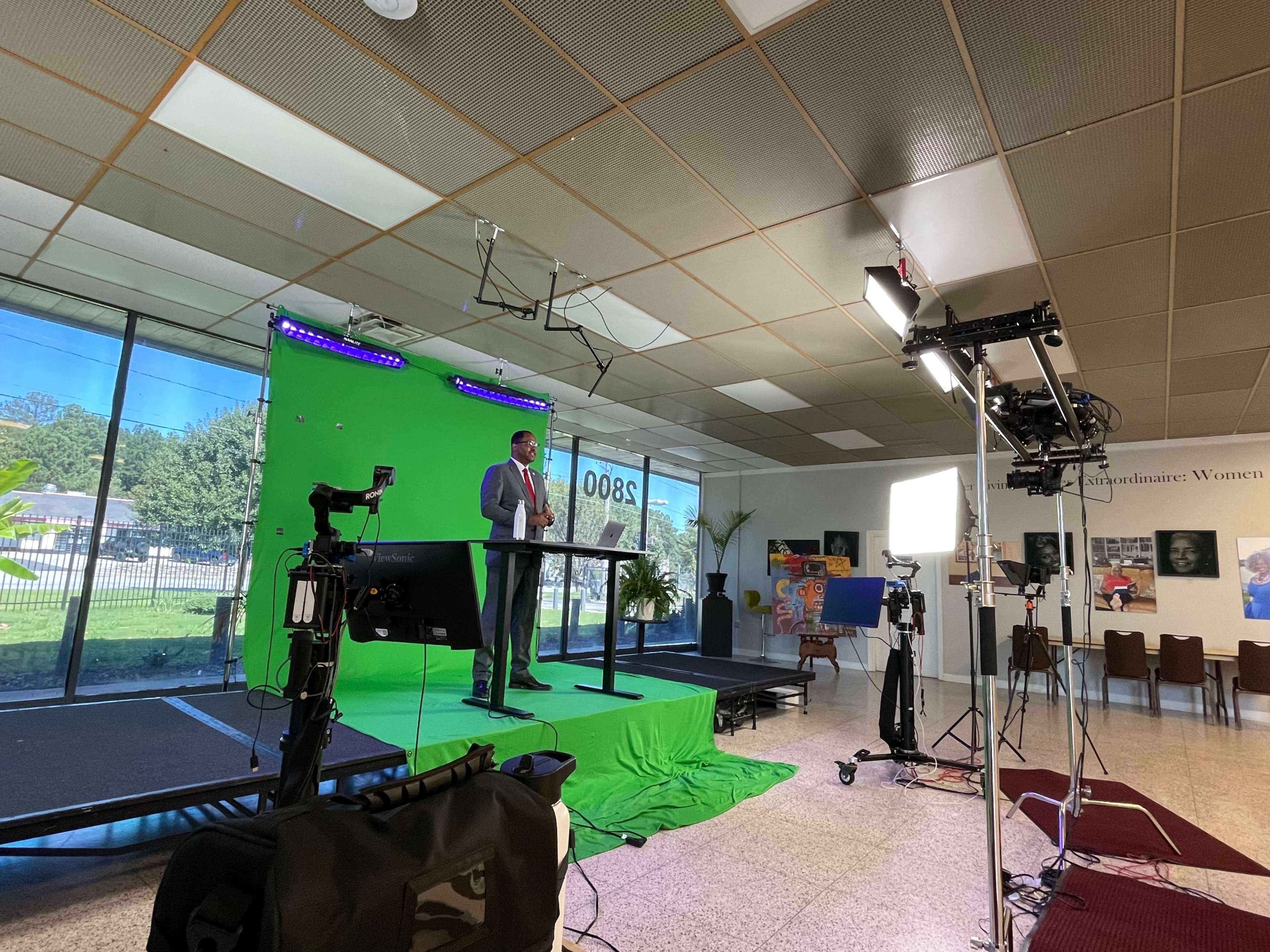 A man stands at a podium in front of a green screen, with cameras and lighting equipment set up in a spacious indoor area.