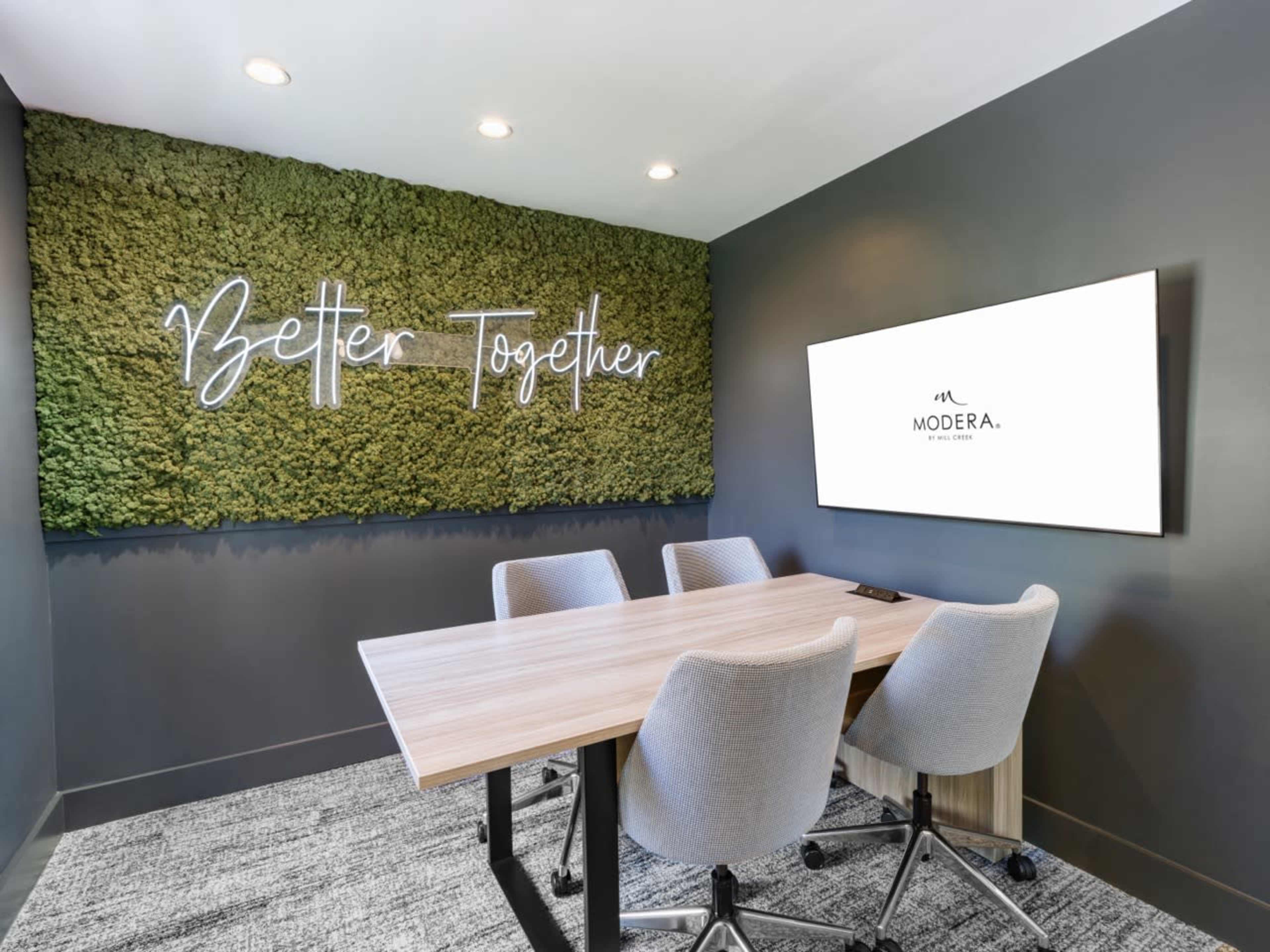 A modern conference room features a moss-covered wall with the phrase "Better Together" illuminated above a sleek table and chairs, along with a mounted display showing the logo of Modera.