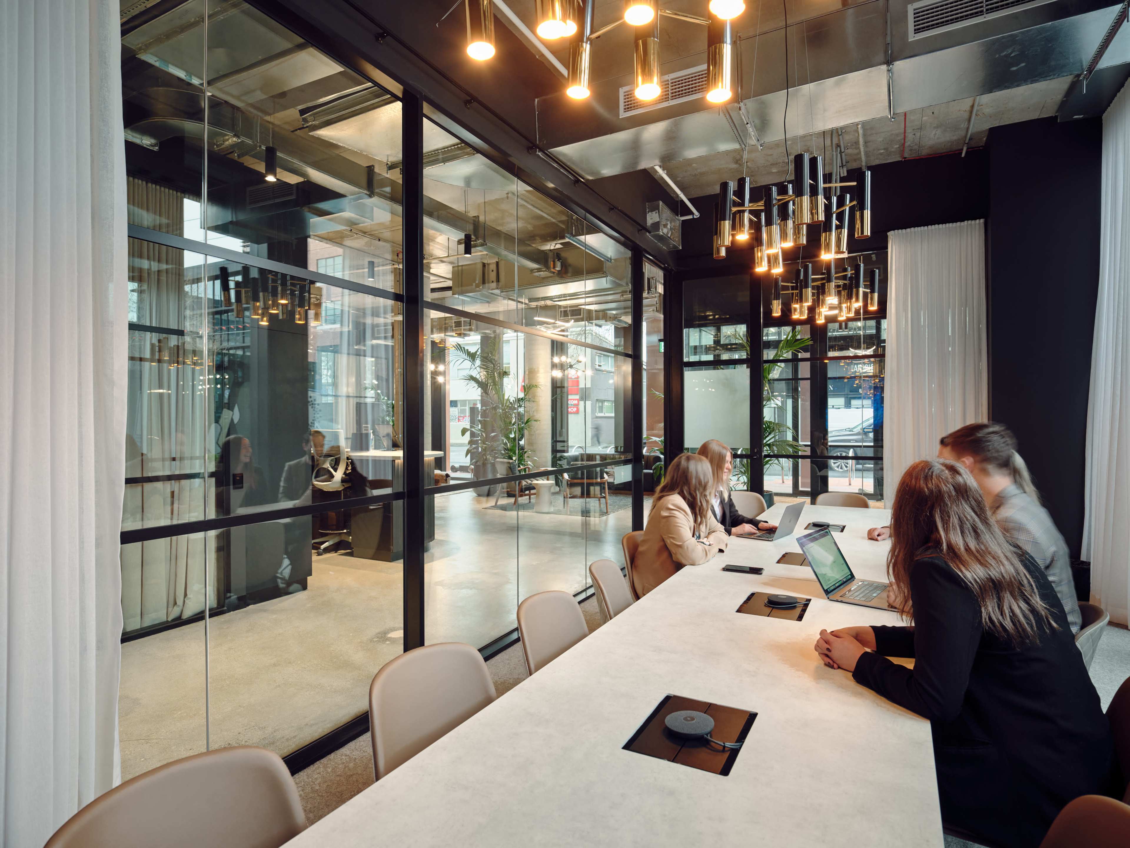 A modern conference room features a long table with laptops, while glass walls allow a view of an adjacent workspace.