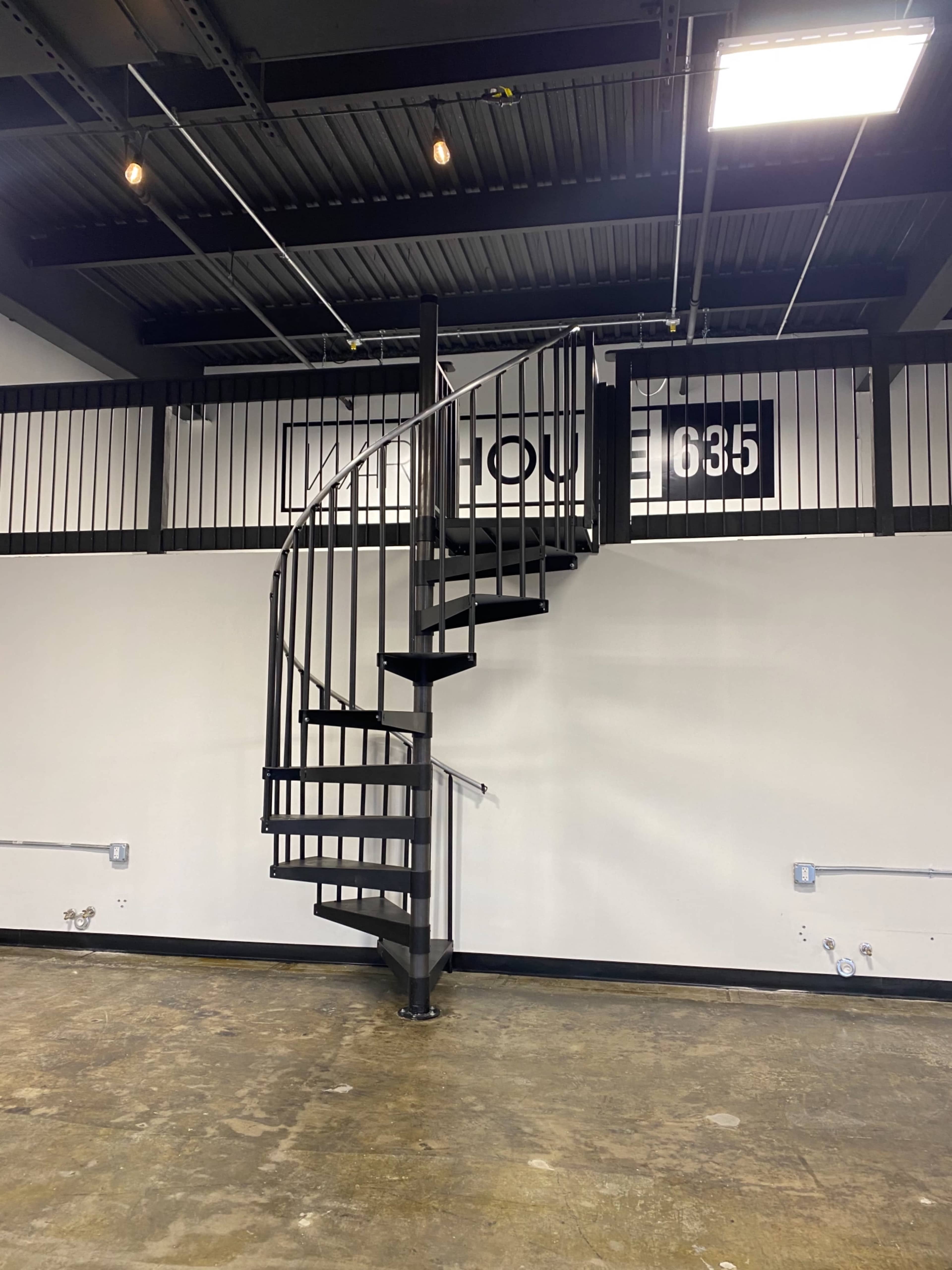 A metal spiral staircase leading to a mezzanine in an industrial-style interior with black railings and concrete flooring.