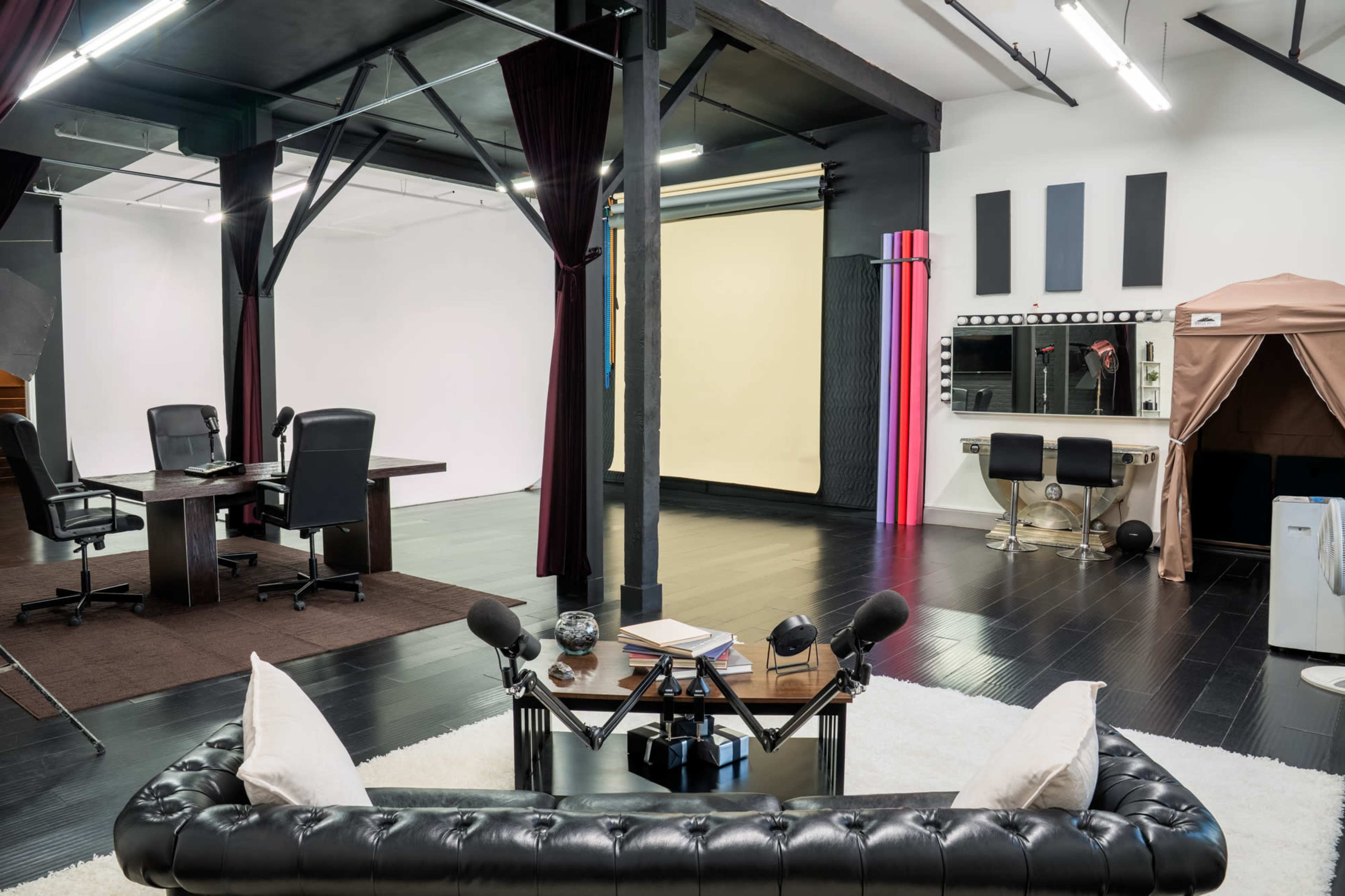 The image shows a spacious studio with a black and white color scheme, featuring a modern seating area, a table with chairs, a backdrop setup, and a vanity area with lights.