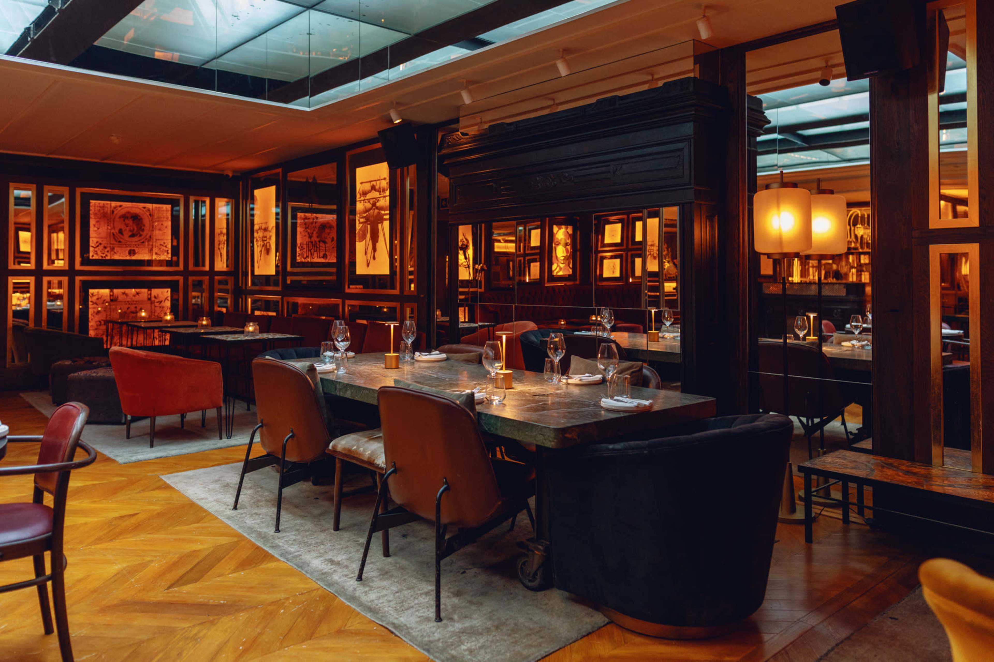 A warmly lit restaurant interior with wooden flooring, framed artwork on the walls, and a mix of seating arrangements including chairs and tables.