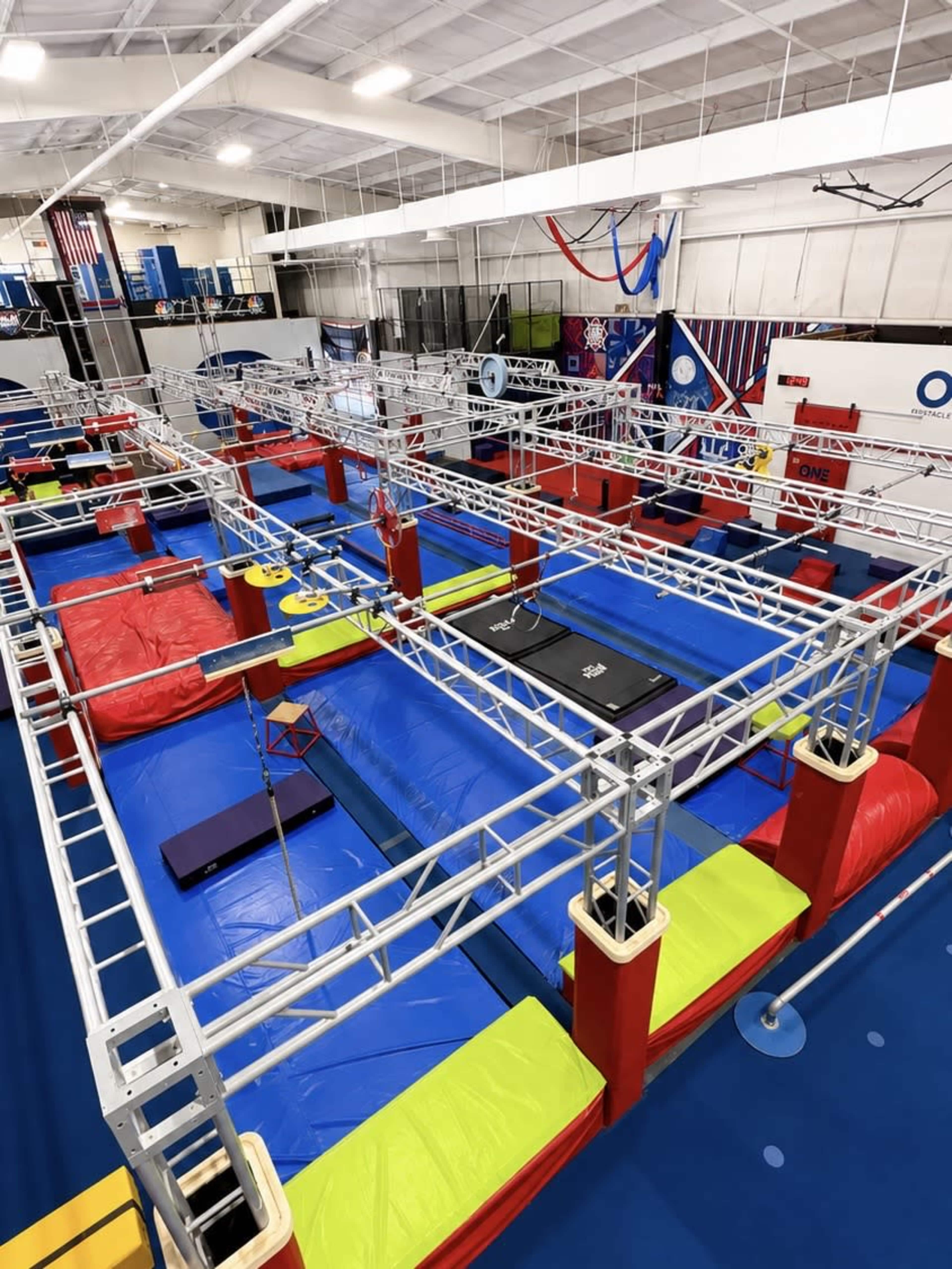 The image shows an indoor obstacle course with various colorful padded platforms, bars, and nets arranged in a large gymnasium.