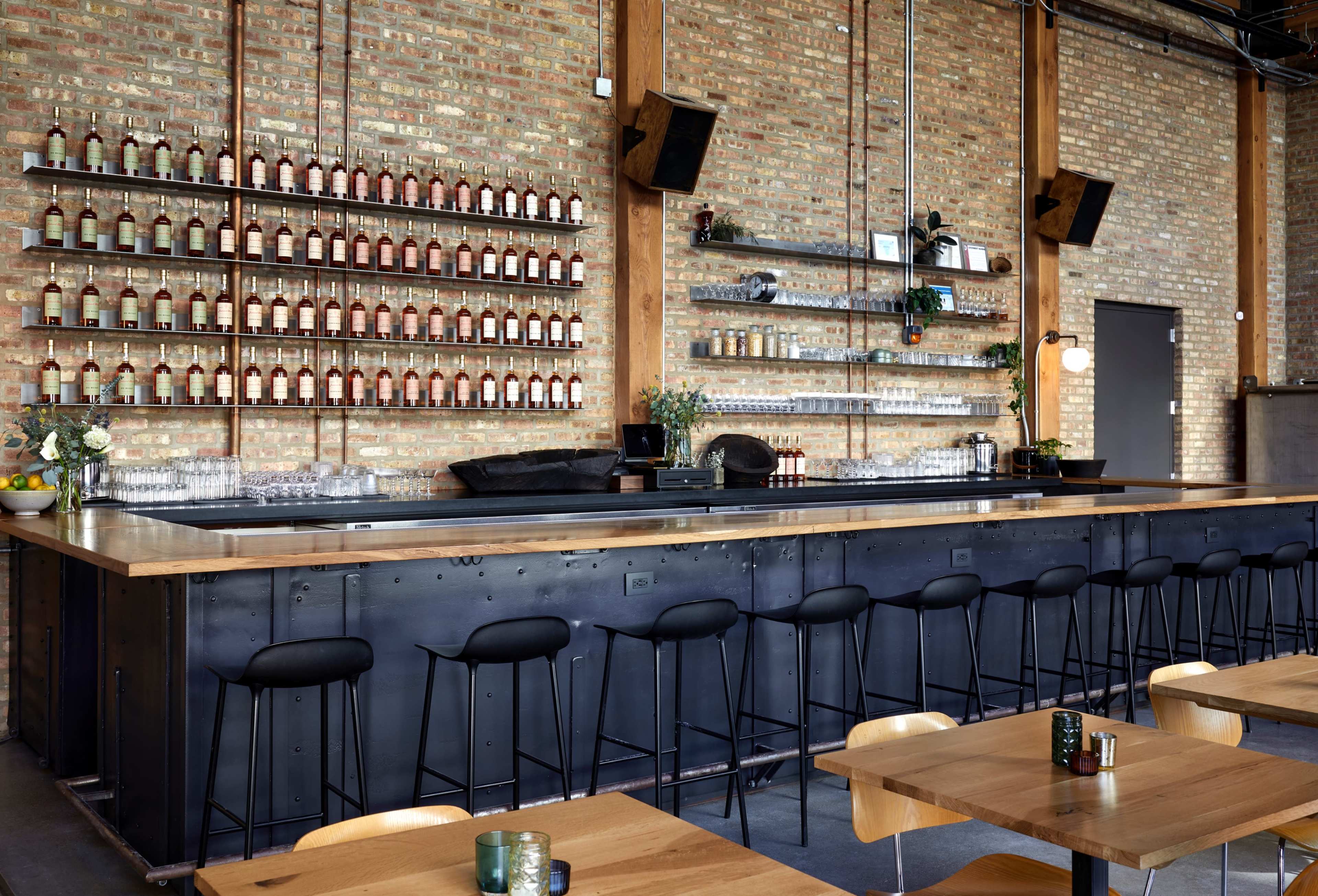 The image shows a modern bar area with a long wooden counter, high black stools, and shelves of bottles against a brick wall.
