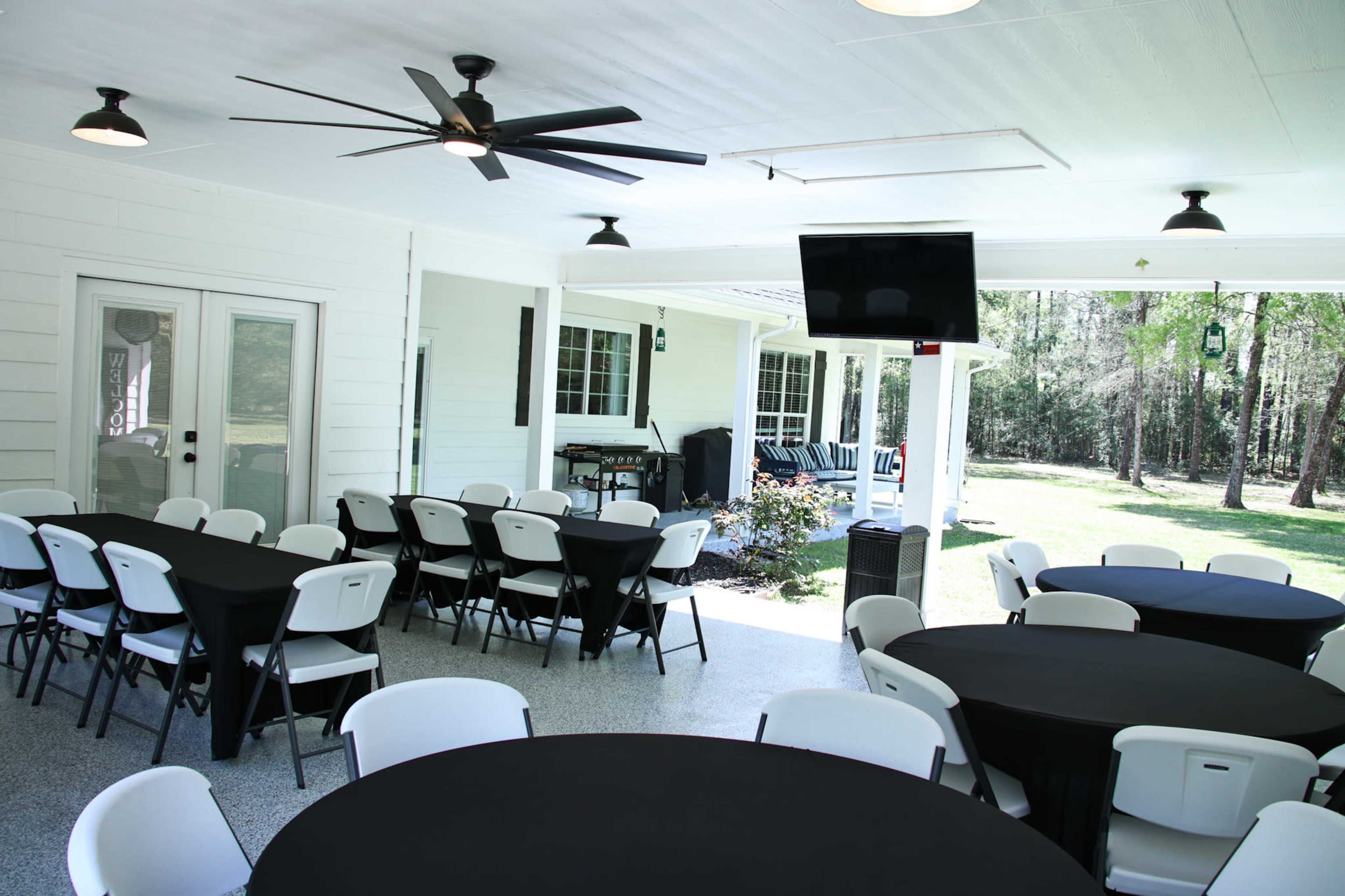 A spacious outdoor room with several round tables covered in black cloth, a ceiling fan, and a television mounted on the wall.