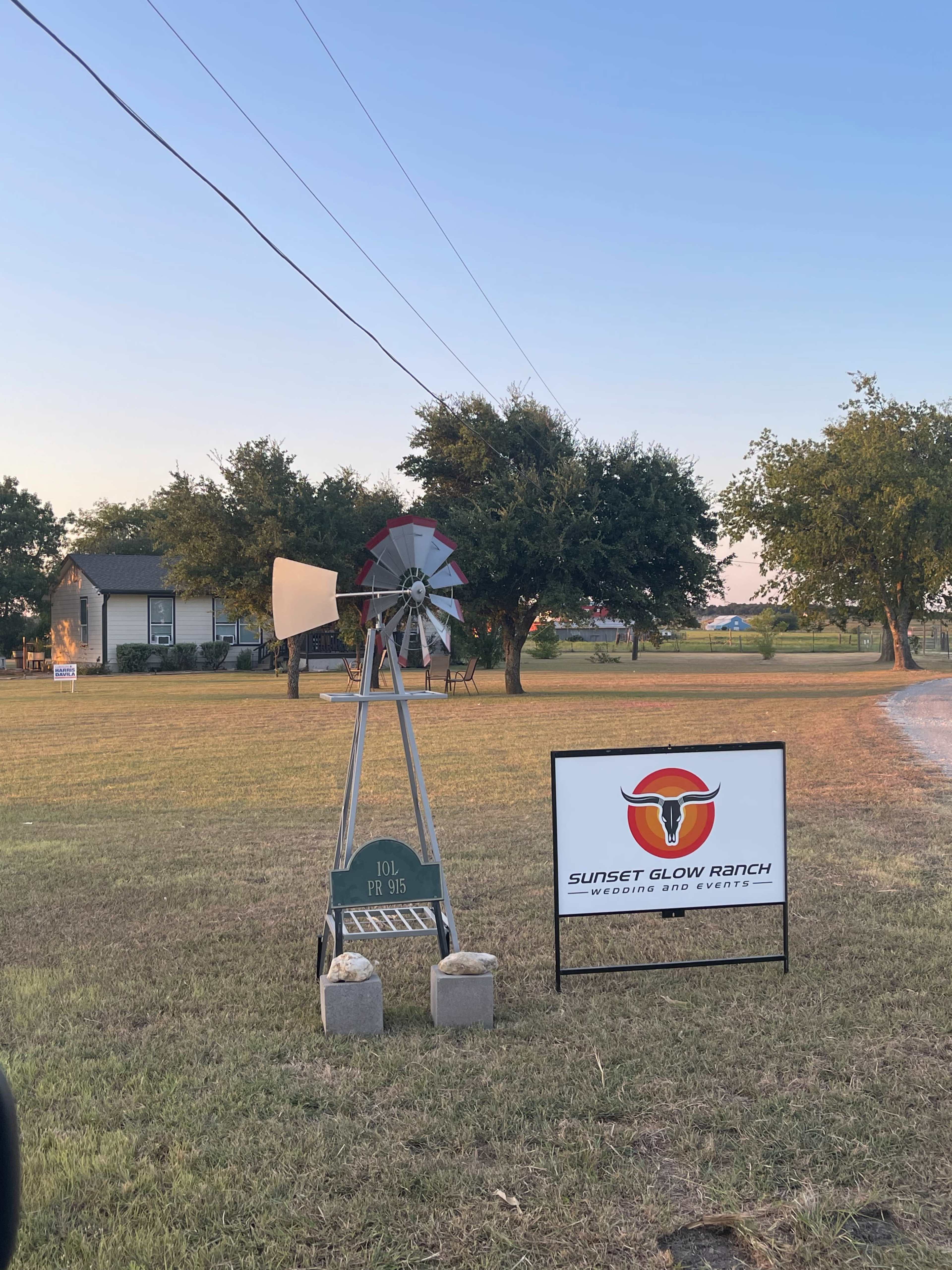 A windmill stands beside a sign for Sunset Glow Ranch in a grassy area near a house and trees.
