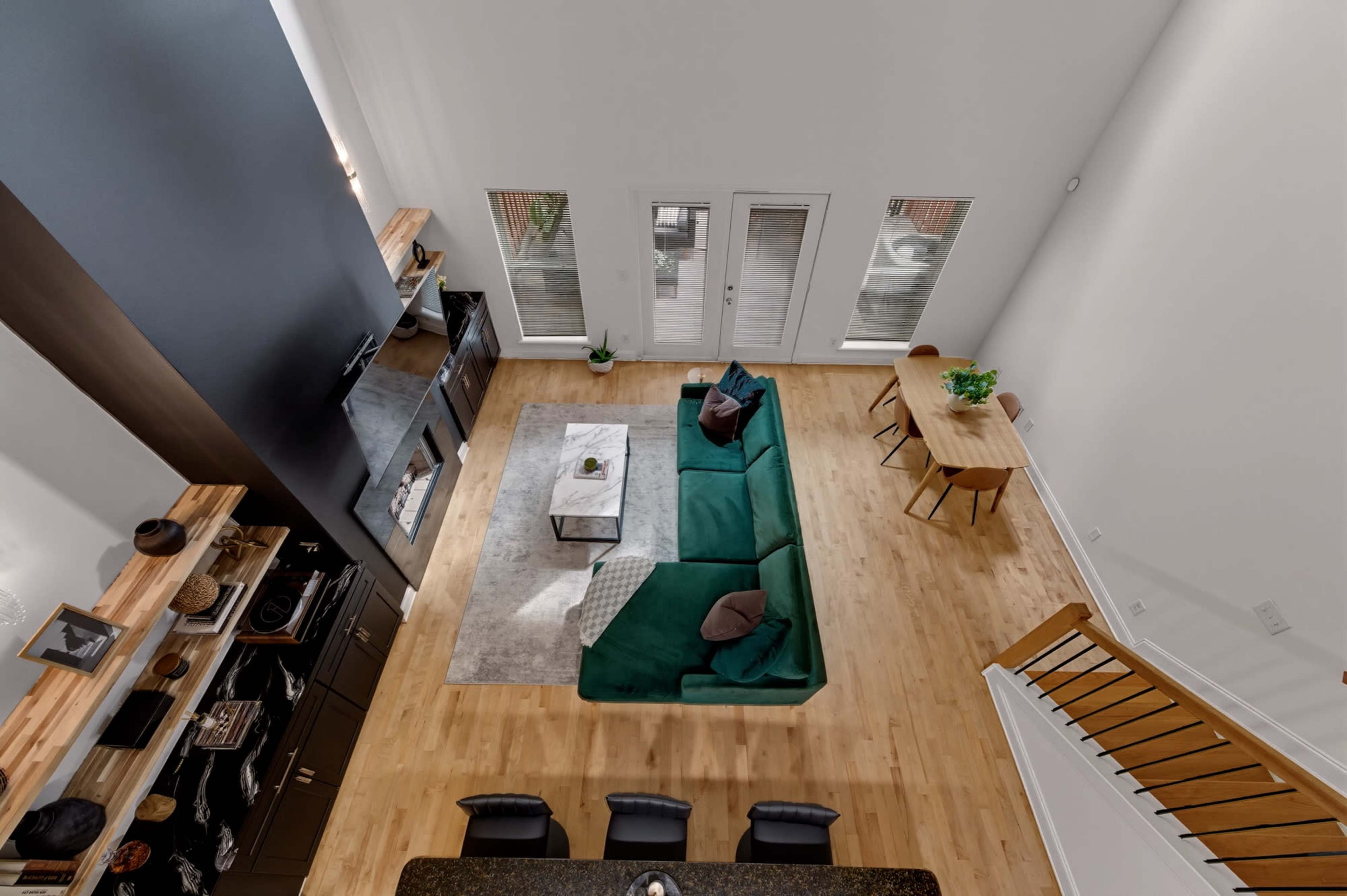 The image shows an aerial view of a spacious living room and dining area featuring a green sofa, a wooden dining table, and large windows.