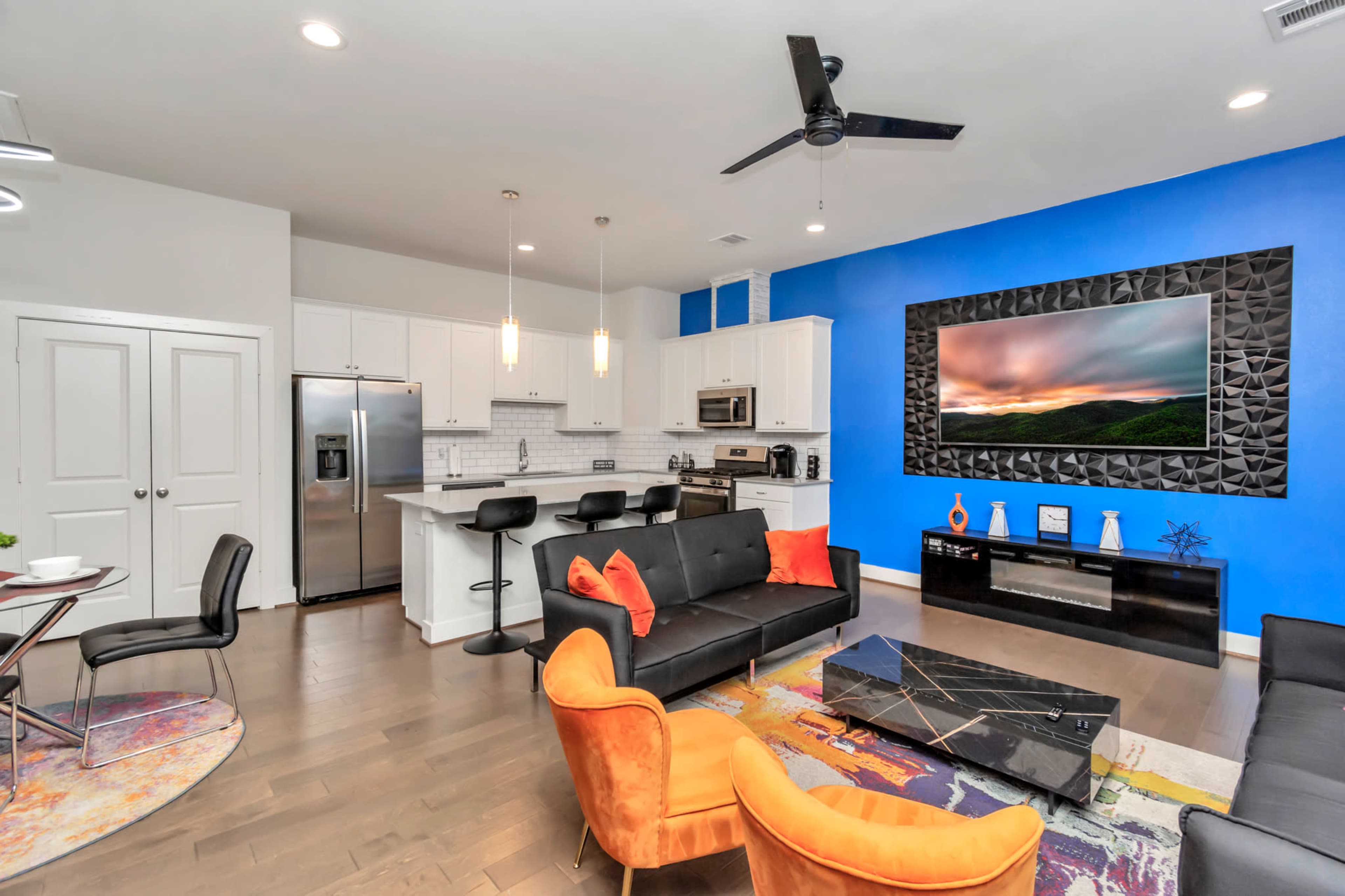 The image shows a modern living space with a black sectional sofa, orange accent chairs, a kitchen area with white cabinets, and a large blue wall featuring a framed landscape picture.