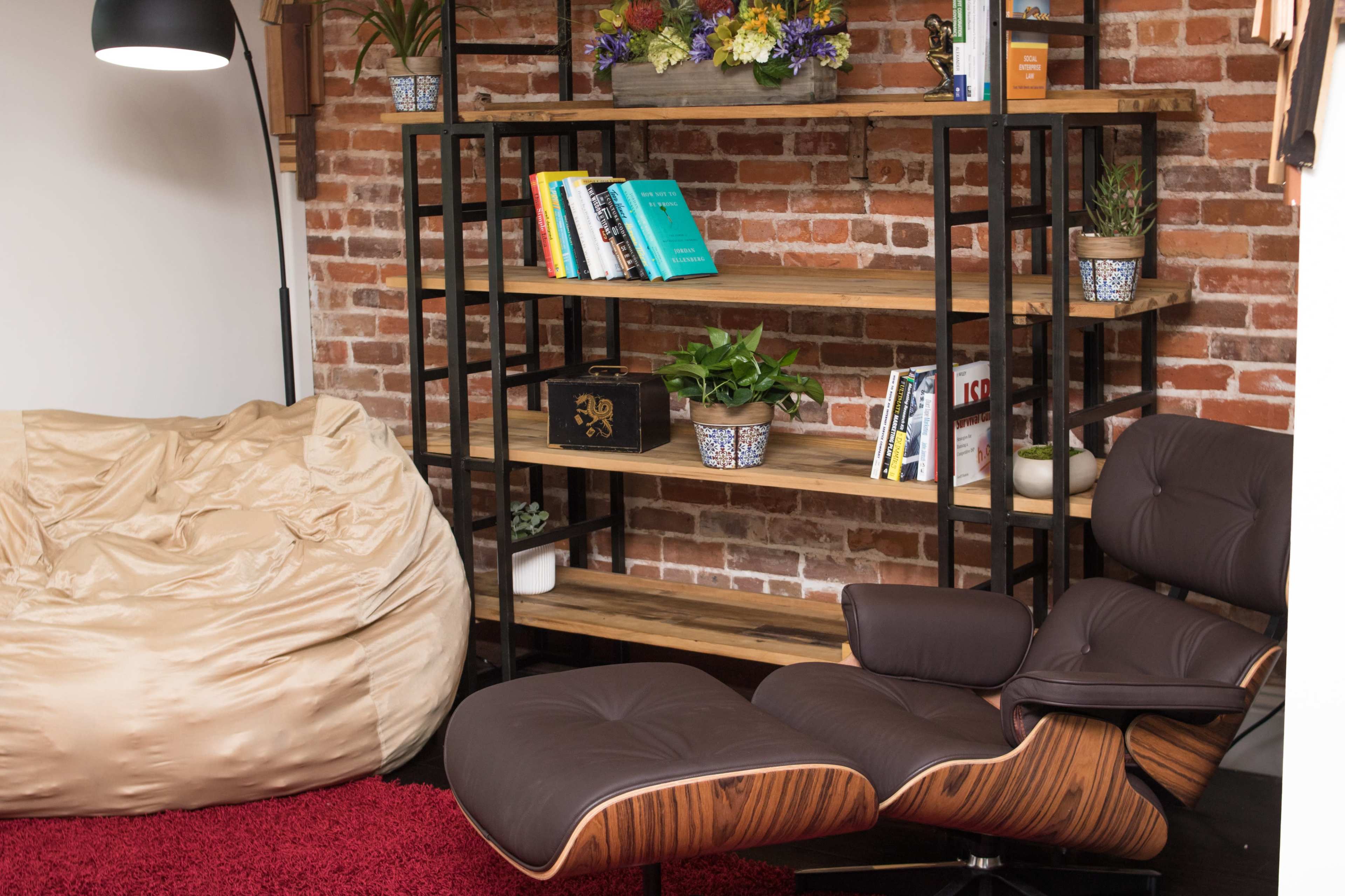 The image shows a cozy room featuring a brown leather chair next to a bookshelf filled with books and plants, with a tan bean bag on one side and a red rug on the floor.