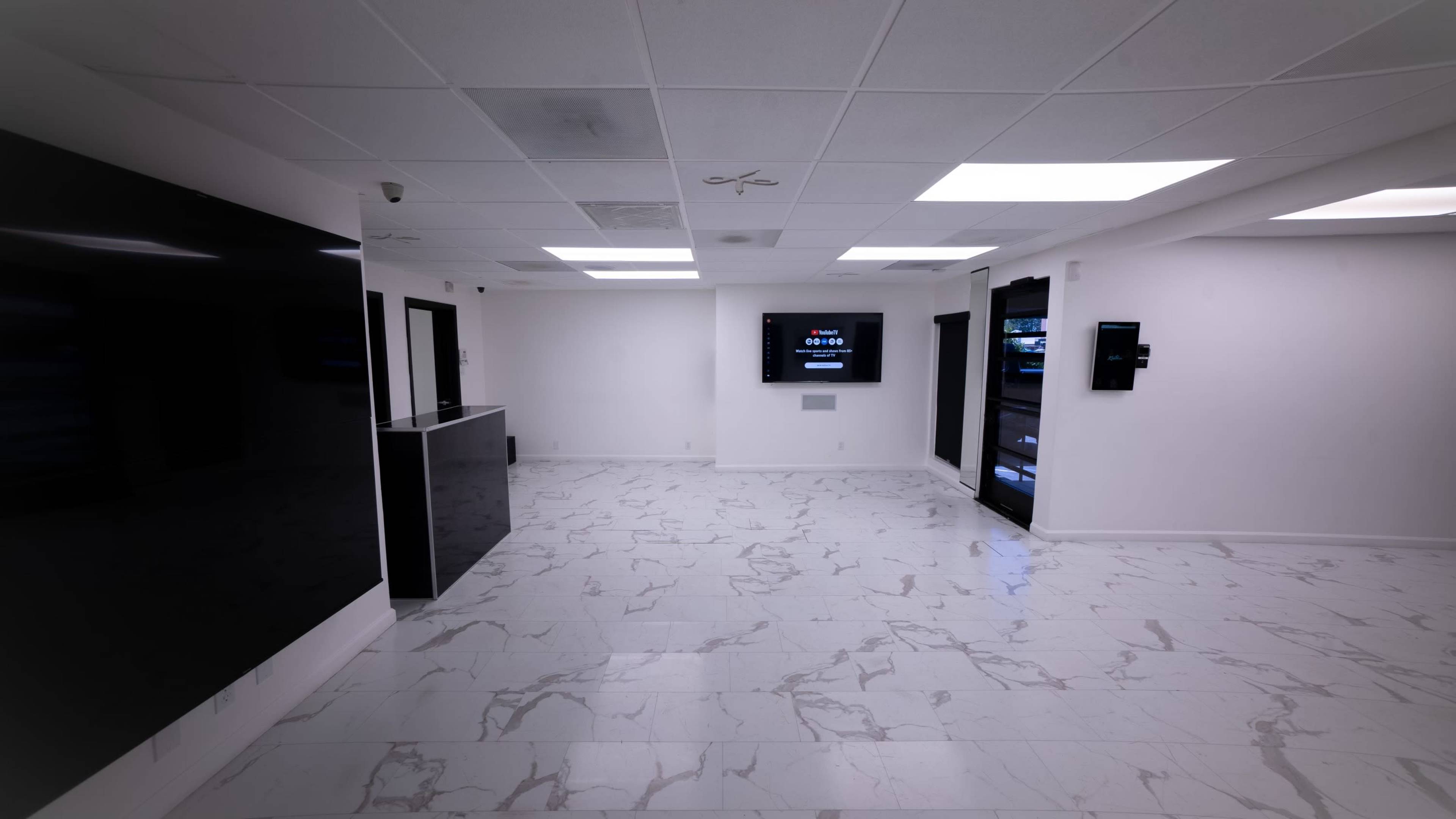 The image shows a modern, spacious interior with white marble flooring, a reception area with a black counter, and multiple screens mounted on the walls.
