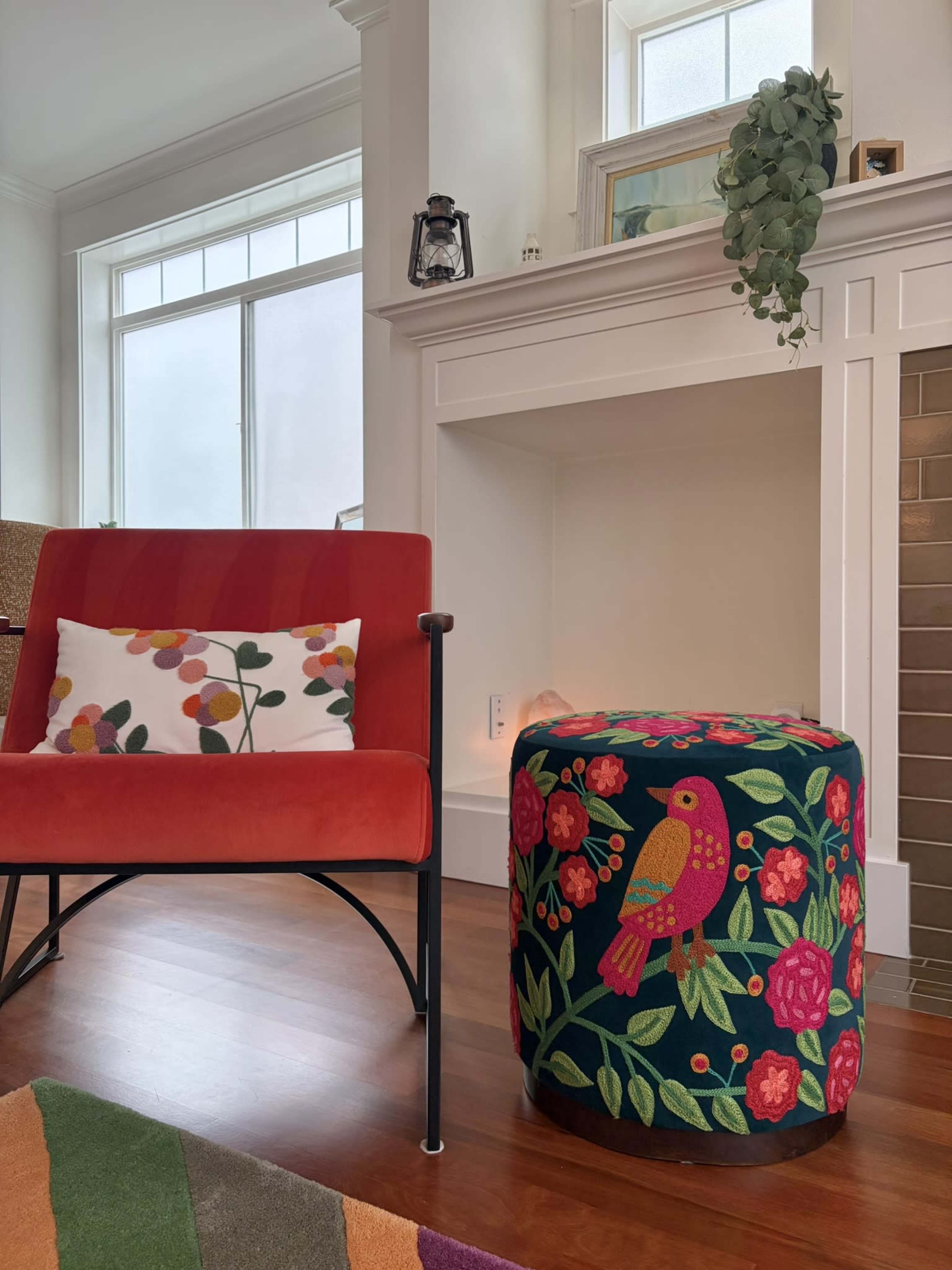 A red chair with a floral pillow sits next to a colorful ottoman adorned with a bird and flowers, near a white fireplace in a well-lit room.