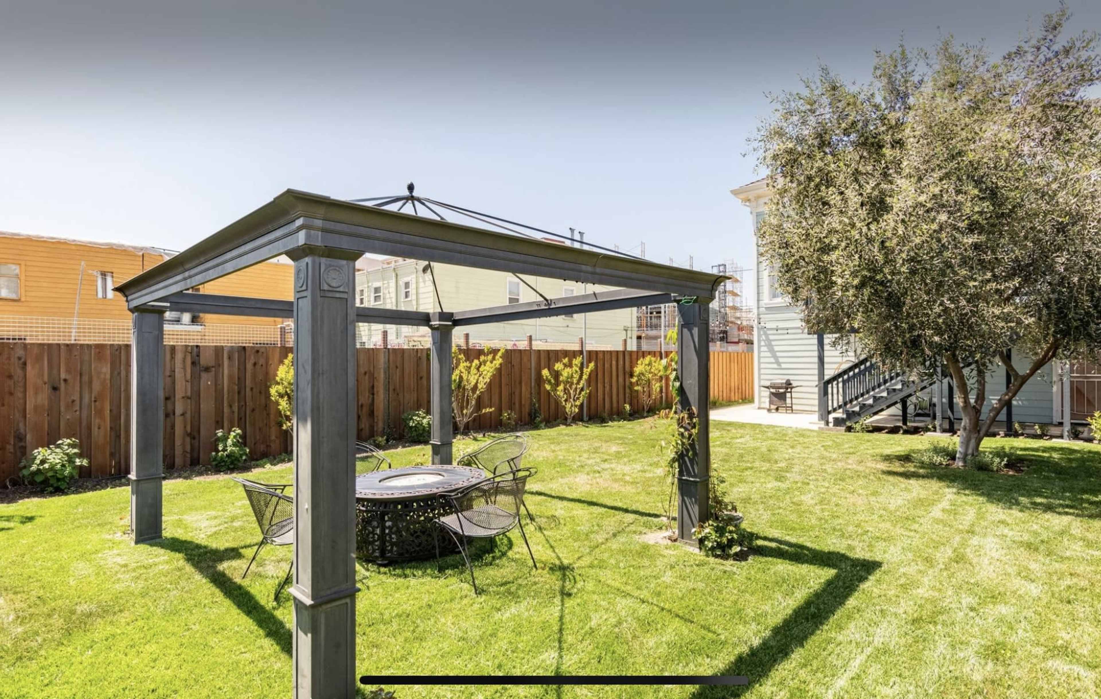 A gazebo with a round table and chairs is situated on a neatly maintained lawn surrounded by a wooden fence and residential buildings.