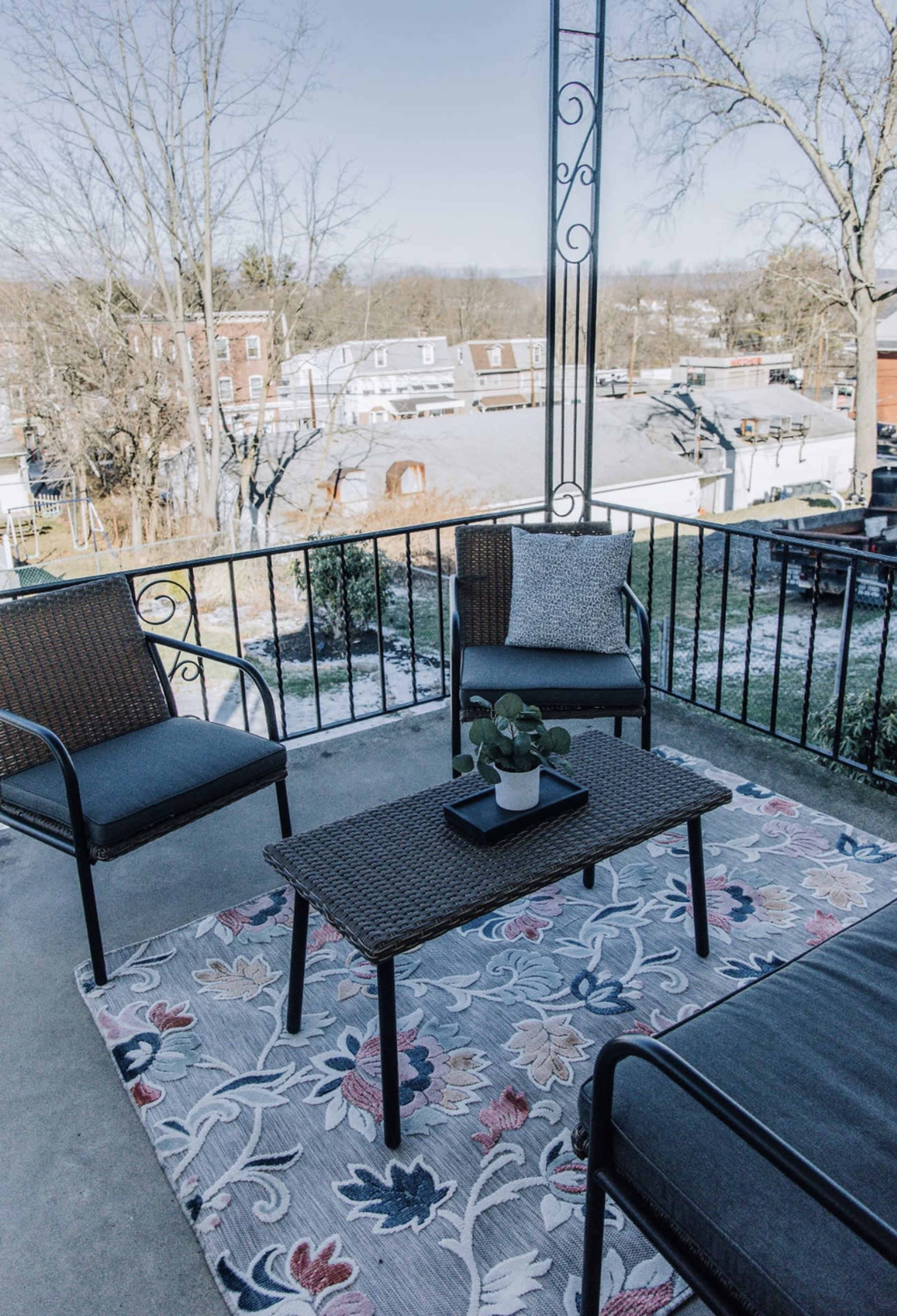 A small outdoor patio area with two chairs and a table on a patterned rug, overlooking a residential neighborhood.