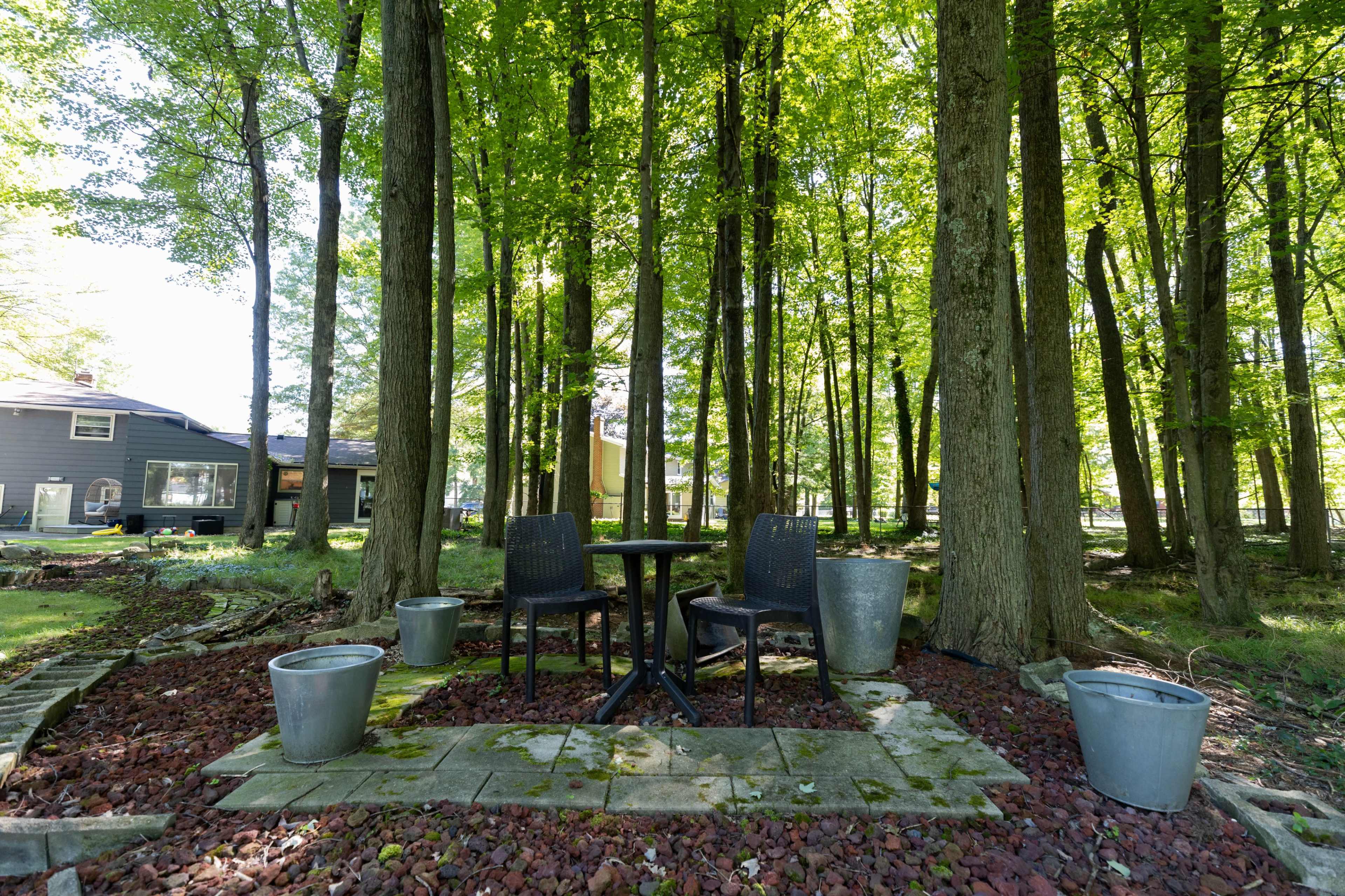 A small seating area with two chairs and a table is surrounded by trees and gravel in a backyard.