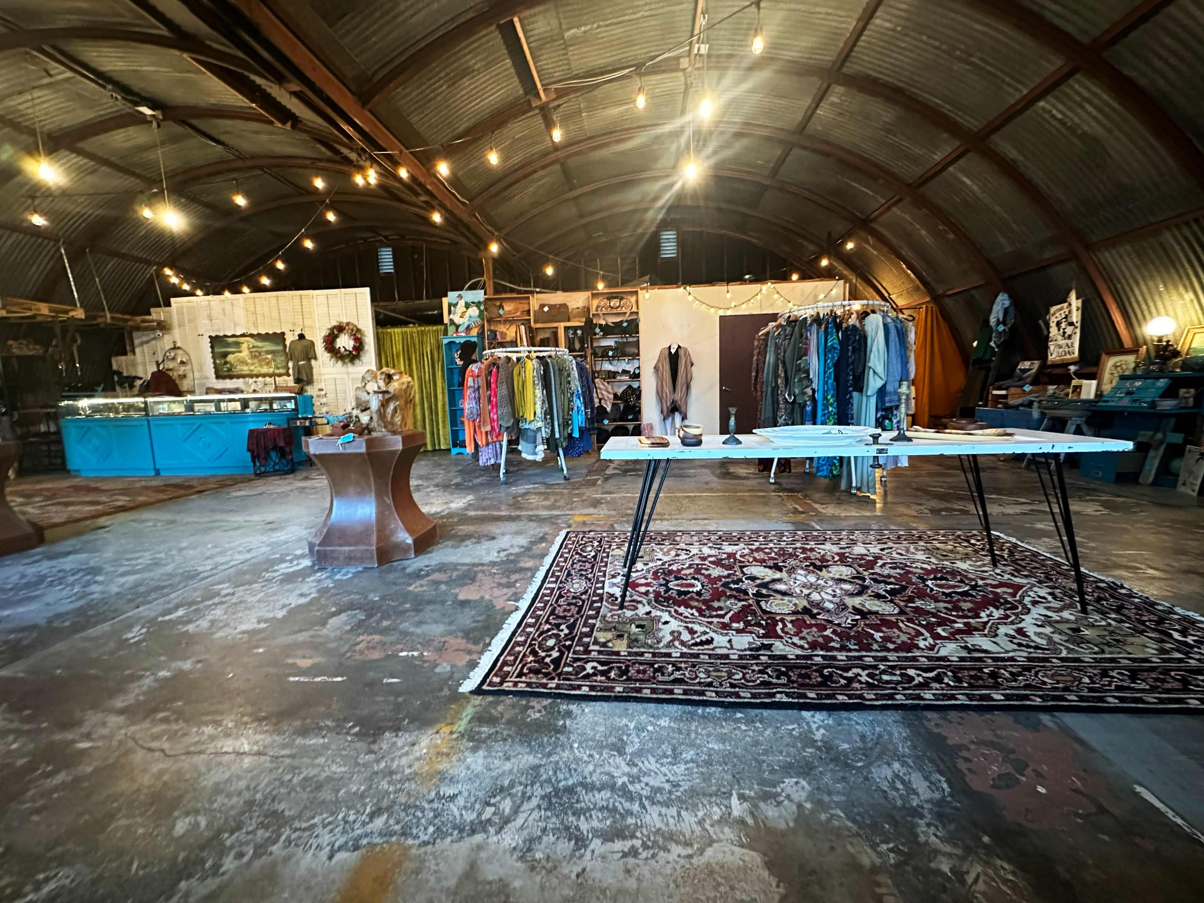 A spacious, rustic retail space with clothing racks and displays arranged around a central table on a concrete floor.