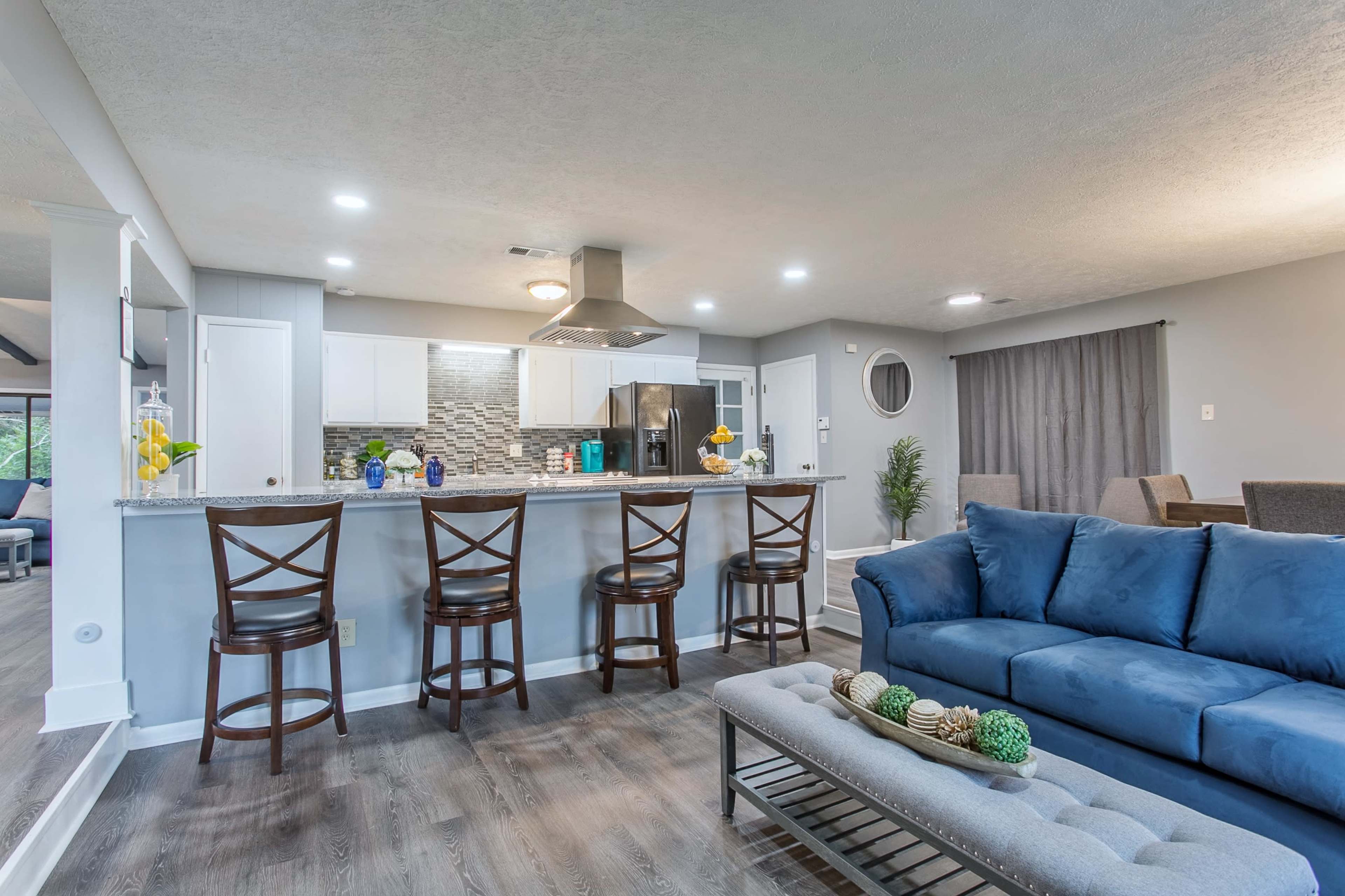 A modern kitchen and living room area featuring a blue sofa, a bar with three stools, and a kitchen island with various decorative items and appliances.