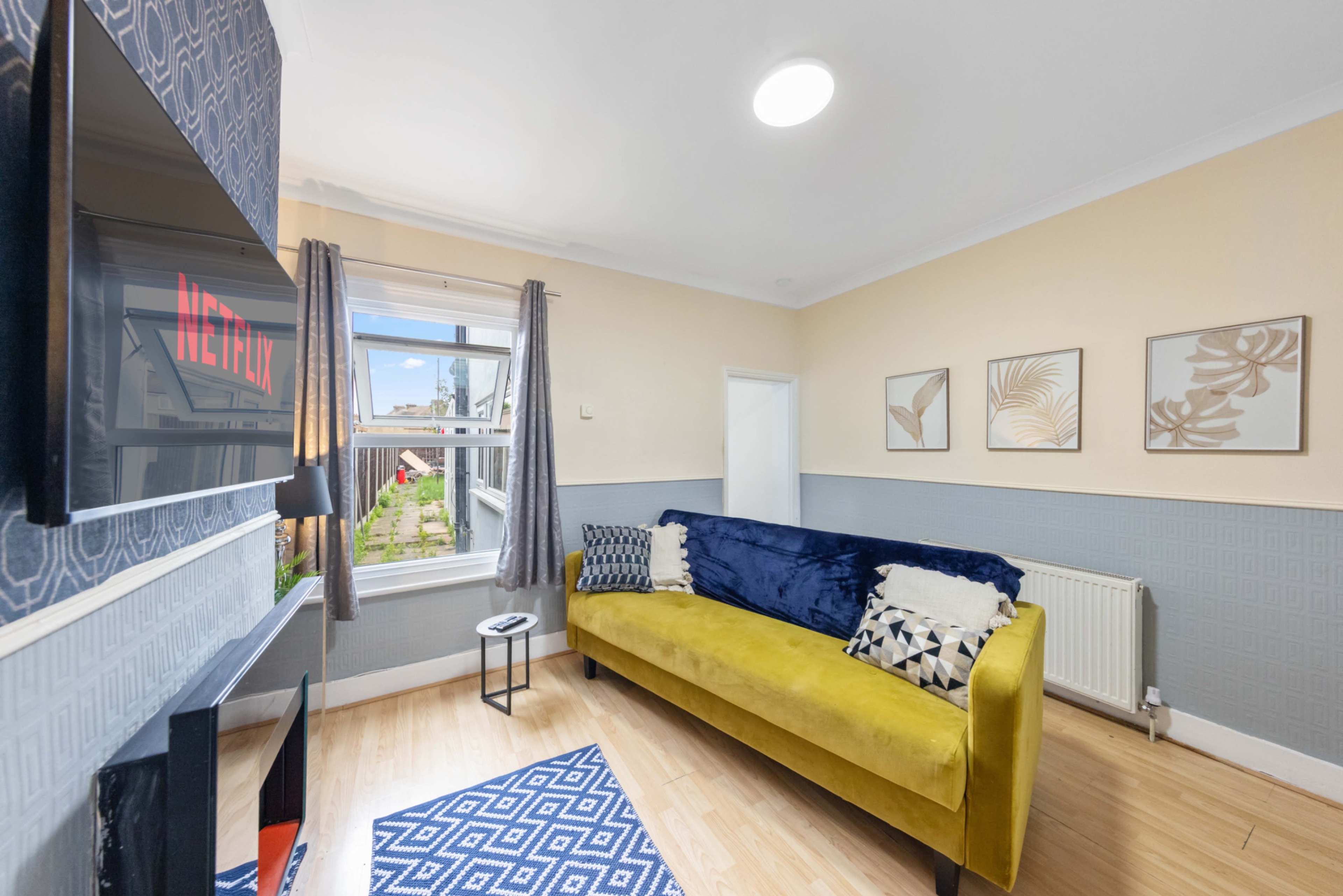 A bright living room features a yellow couch, a black TV mounted on the wall, and framed artwork above a blue carpet.